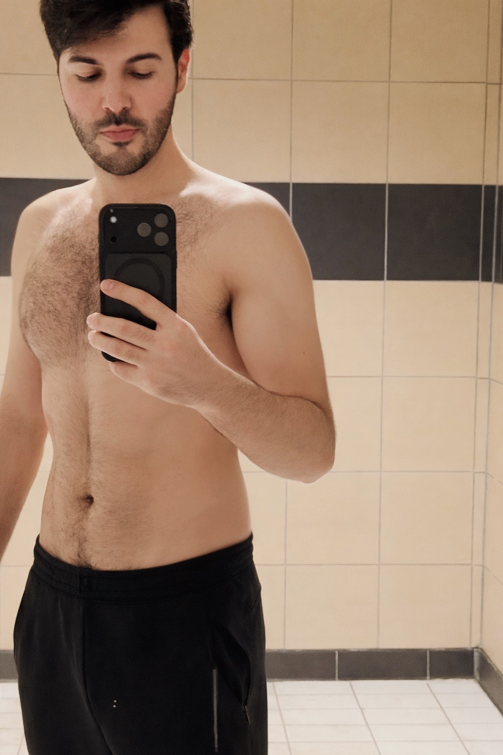 A man takes a mirror selfie in what appears to be a gym locker room. He is shirtless and wearing black sweatpants.