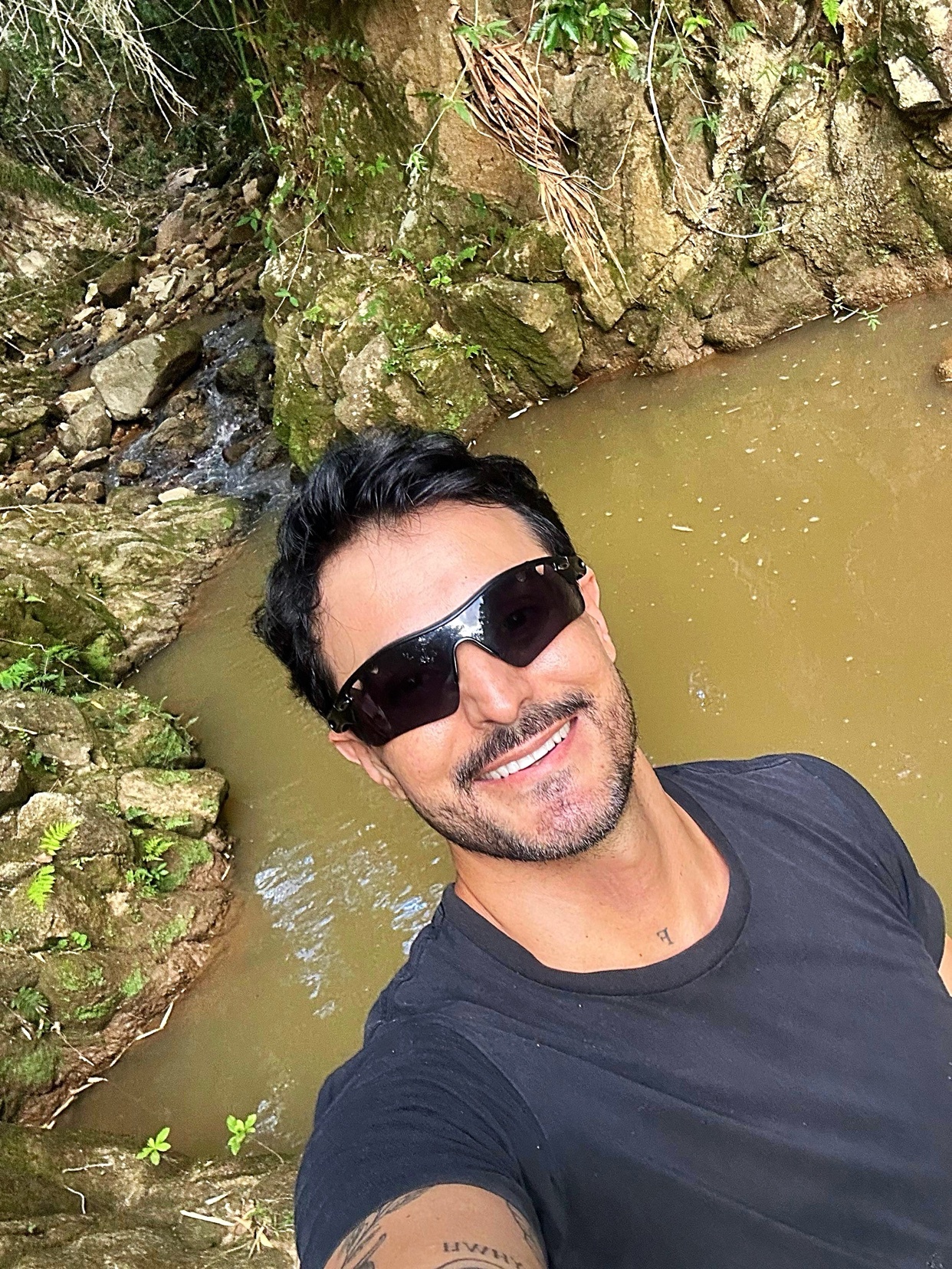 A man with dark hair and stubble smiles at the camera while wearing sunglasses and a black t-shirt. He is taking a selfie outdoors with a natural background of rocks and water.