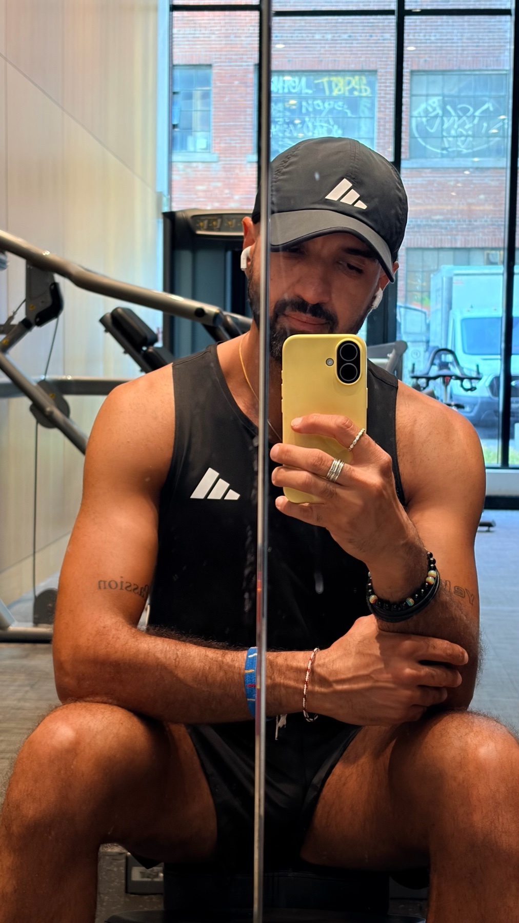 A man in a black tank top and shorts takes a selfie in a gym mirror. He is wearing a baseball cap and earbuds, and has a beard and tattoos.