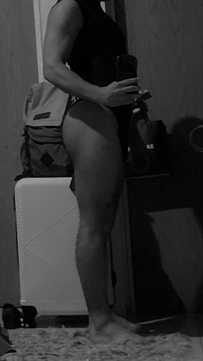 A black and white, side-profile, full-body shot of a man in athletic wear, posing in what appears to be a gym or workout space. The man is showing off his muscular physique, with a focus on his legs and arms.