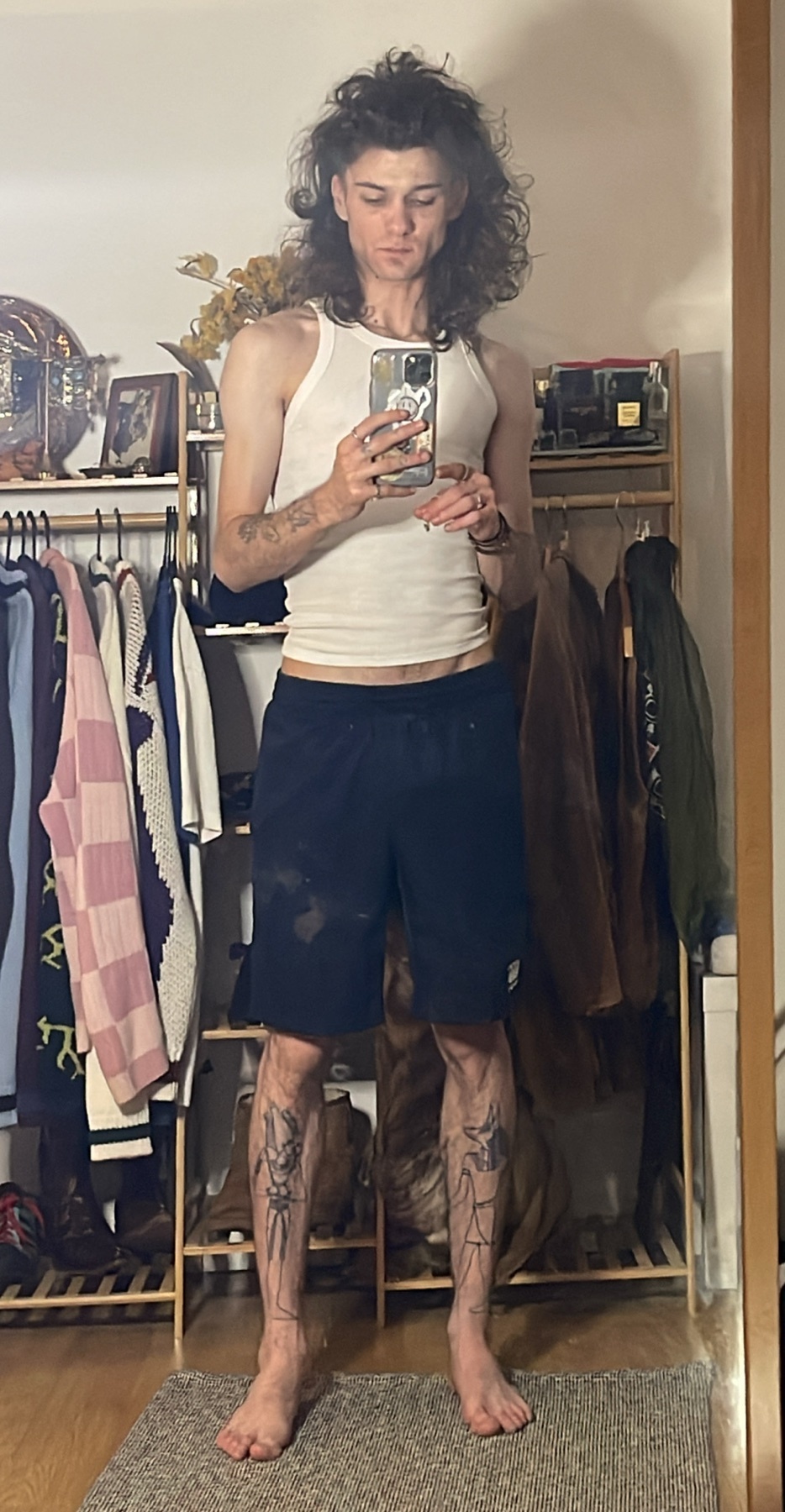 A man takes a mirror selfie, showcasing his physique and tattoos. He's wearing a white tank top and dark shorts.