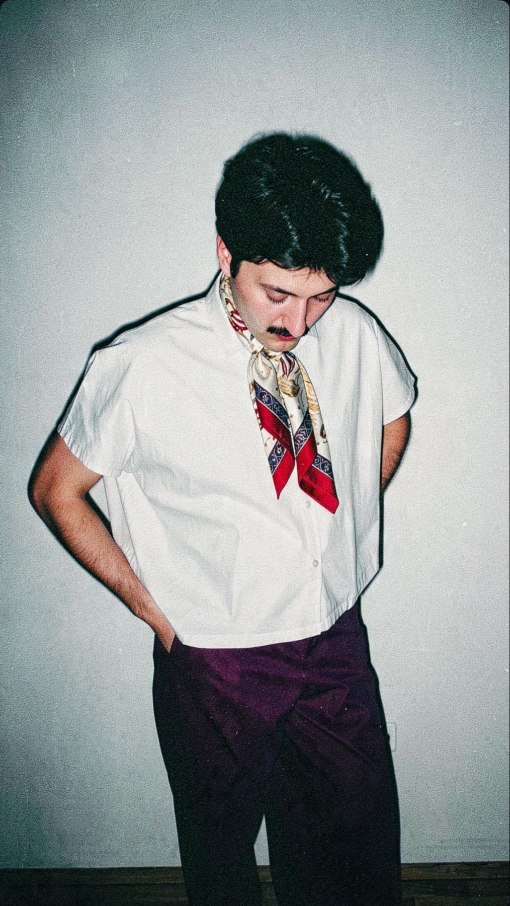 A man with a mustache poses against a plain wall, wearing a white short-sleeved shirt, a colorful scarf tied around his neck, and dark purple pants.