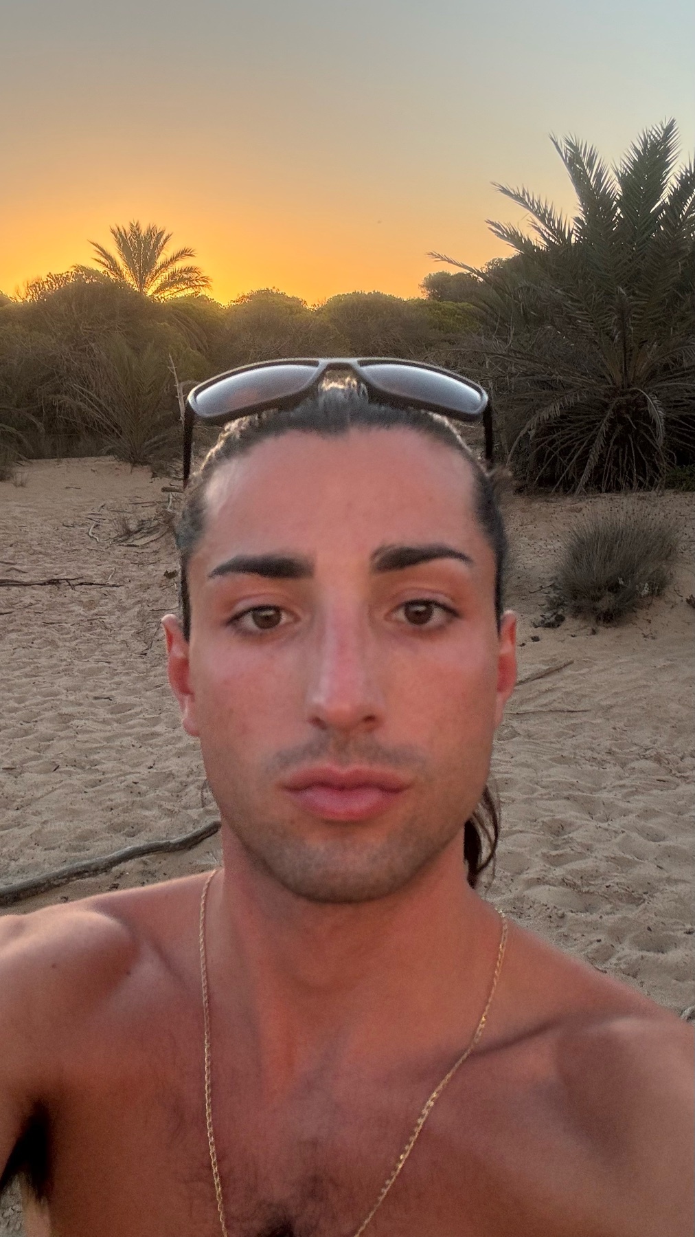A man with long dark hair pulled back and sunglasses perched on his head takes a selfie on a sandy beach at sunset. Palm trees and brush are visible in the background.