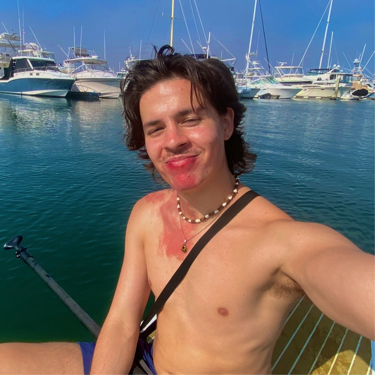 A young man with dark, wavy hair smiles at the camera while sitting on a paddleboard. He is shirtless, wearing a necklace, and has a red mark on his chest. In the background, several boats are docked in a marina under a clear blue sky.
