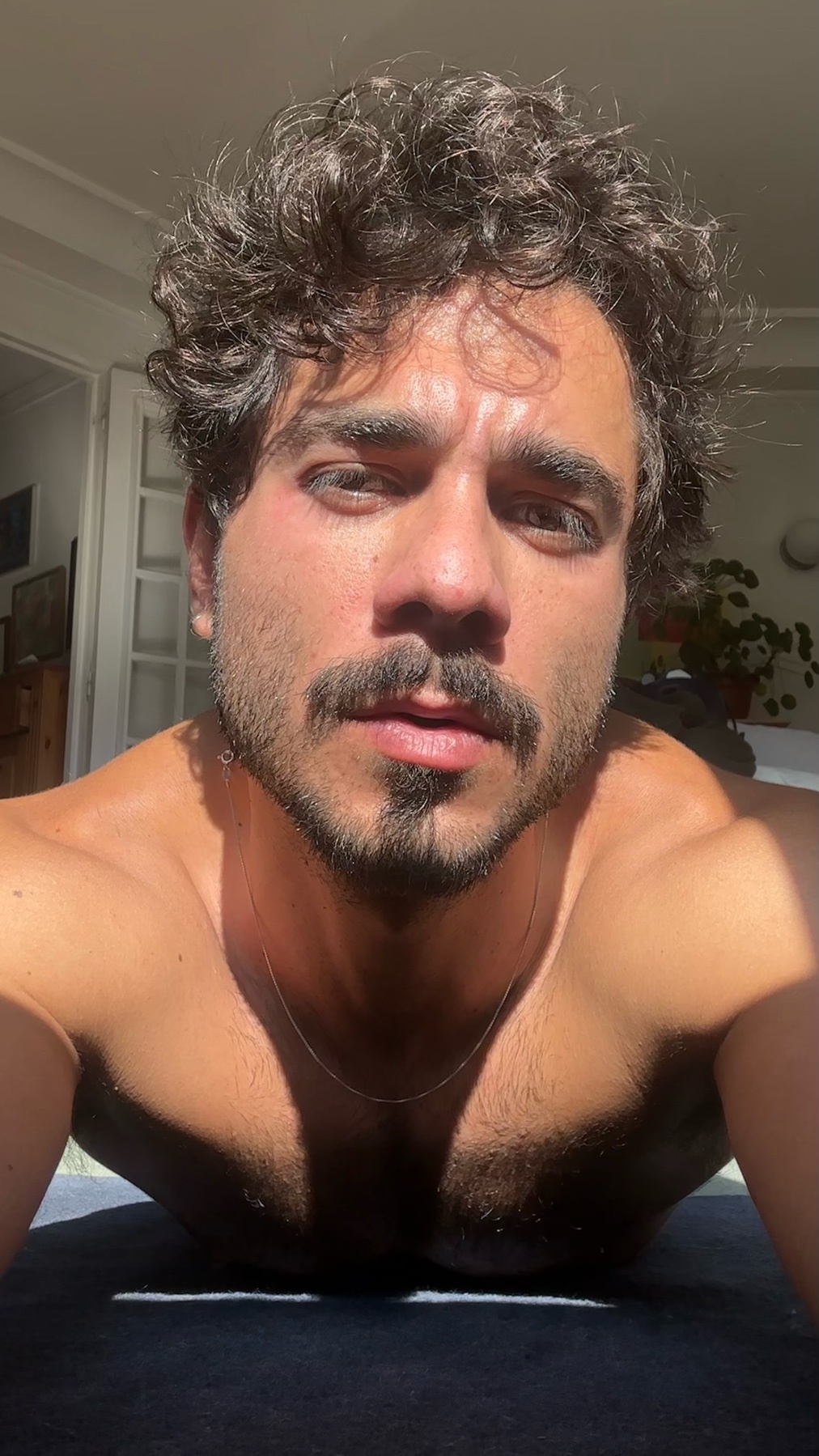 A shirtless man with curly dark hair and a moustache poses for a selfie in natural light.