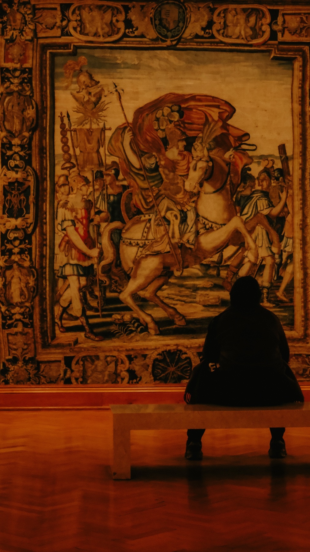 A person sits on a bench in front of a large, ornate tapestry depicting a historical scene with a figure on horseback. The artwork is displayed in a dimly lit gallery.