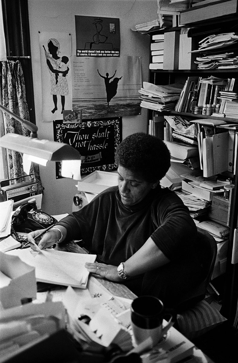 A black and white portrait of Audre Lorde at her desk. She is surrounded by books and papers.