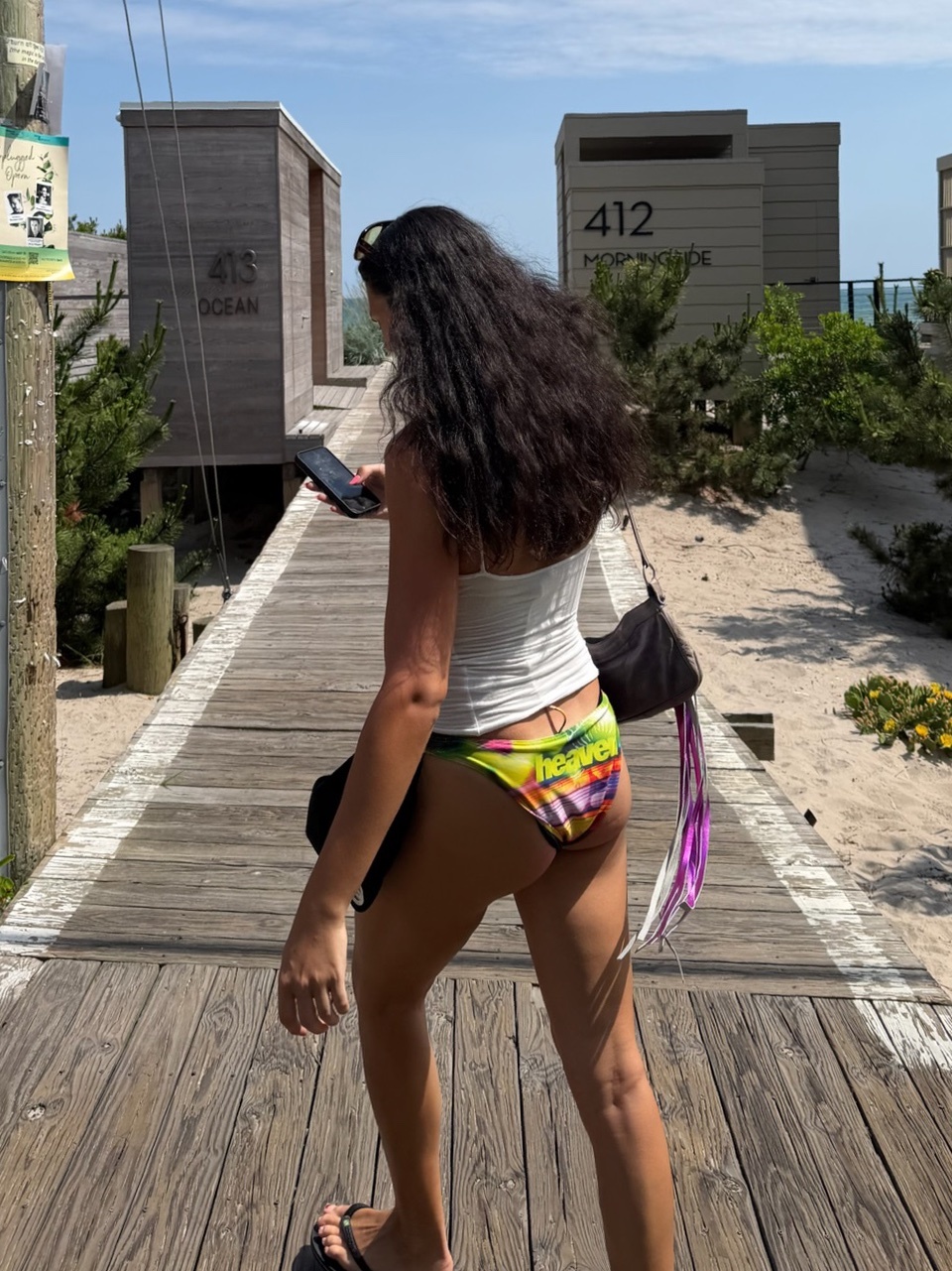 A woman in a bikini walks on a wooden boardwalk towards the beach, holding her phone. The sun shines brightly on this summer day.