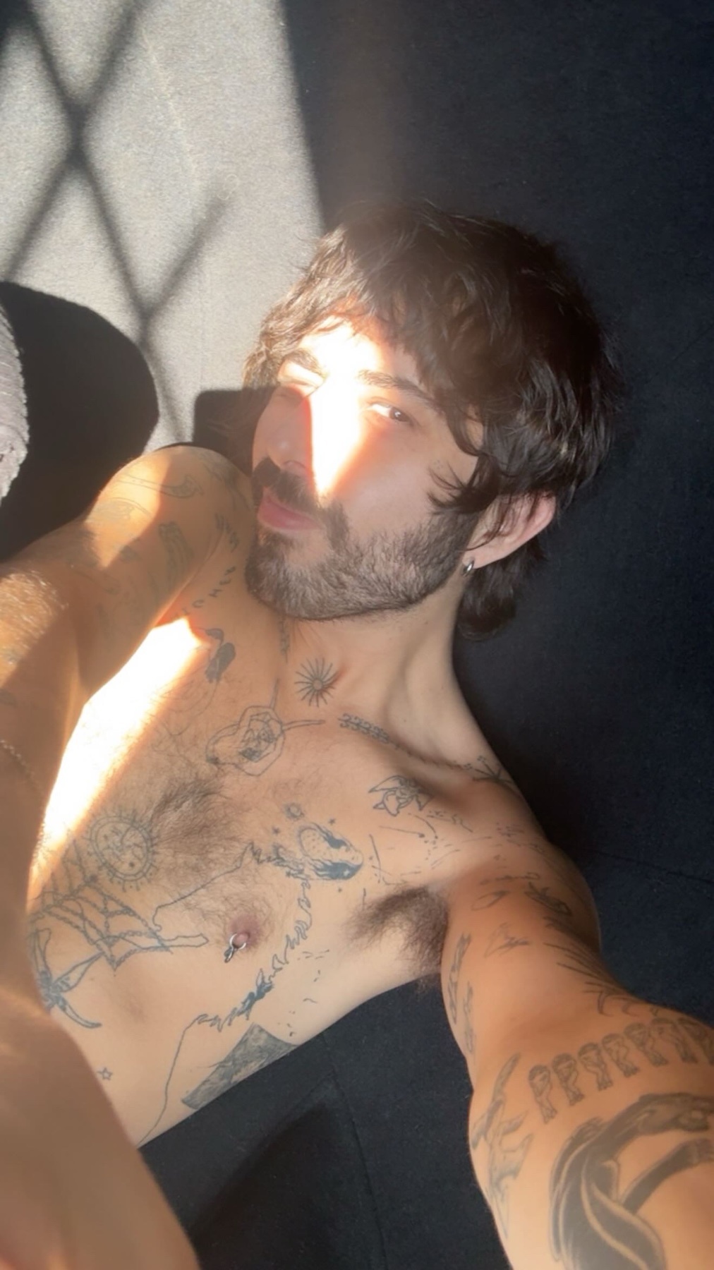 A shirtless man with tattoos poses for a selfie in the sunlight. The image highlights his tattoos and facial hair.
