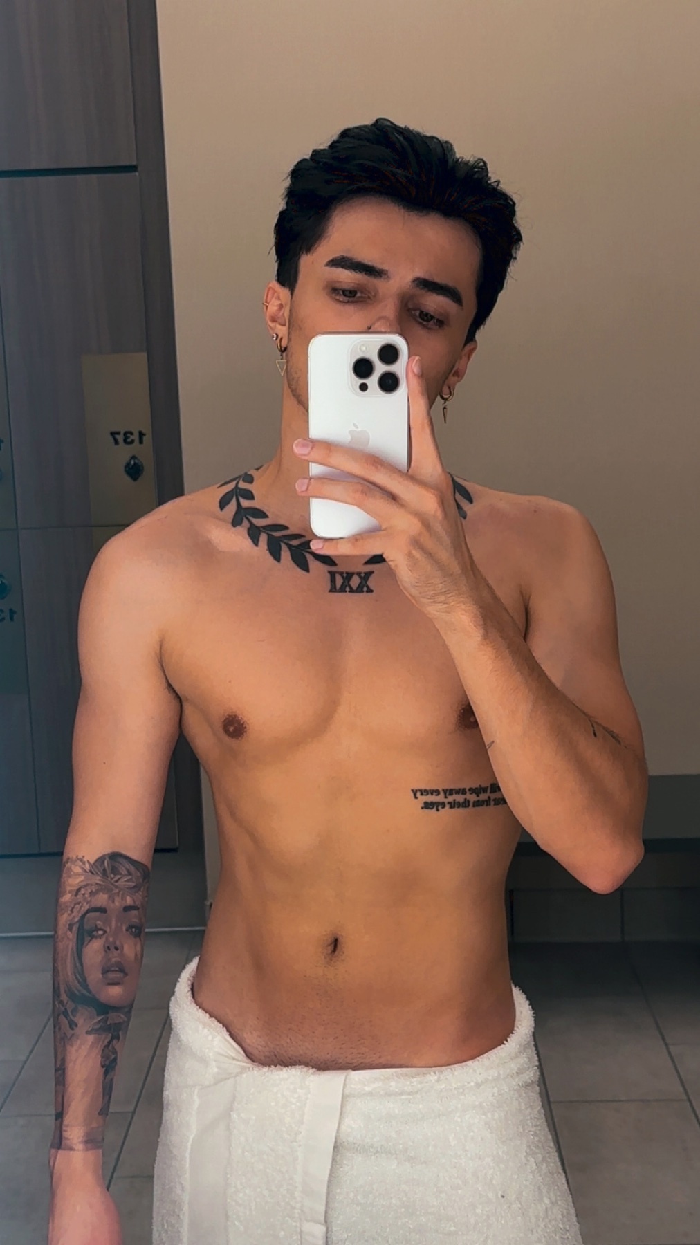 A young man with dark hair and tattoos poses for a selfie while wrapped in a towel.