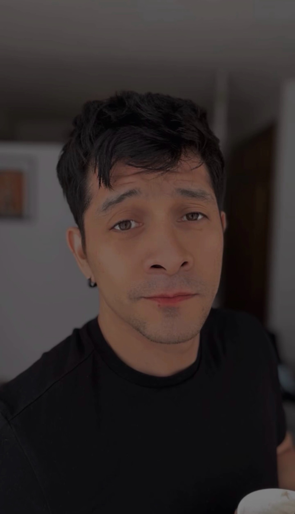 A close-up selfie of a man with dark, wavy hair and a black t-shirt. He is looking directly at the camera with a neutral expression.