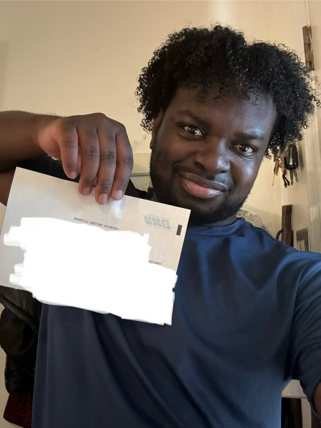 A man with curly dark hair and a beard smiles as he holds up a license to the camera. He is wearing a dark blue t-shirt.