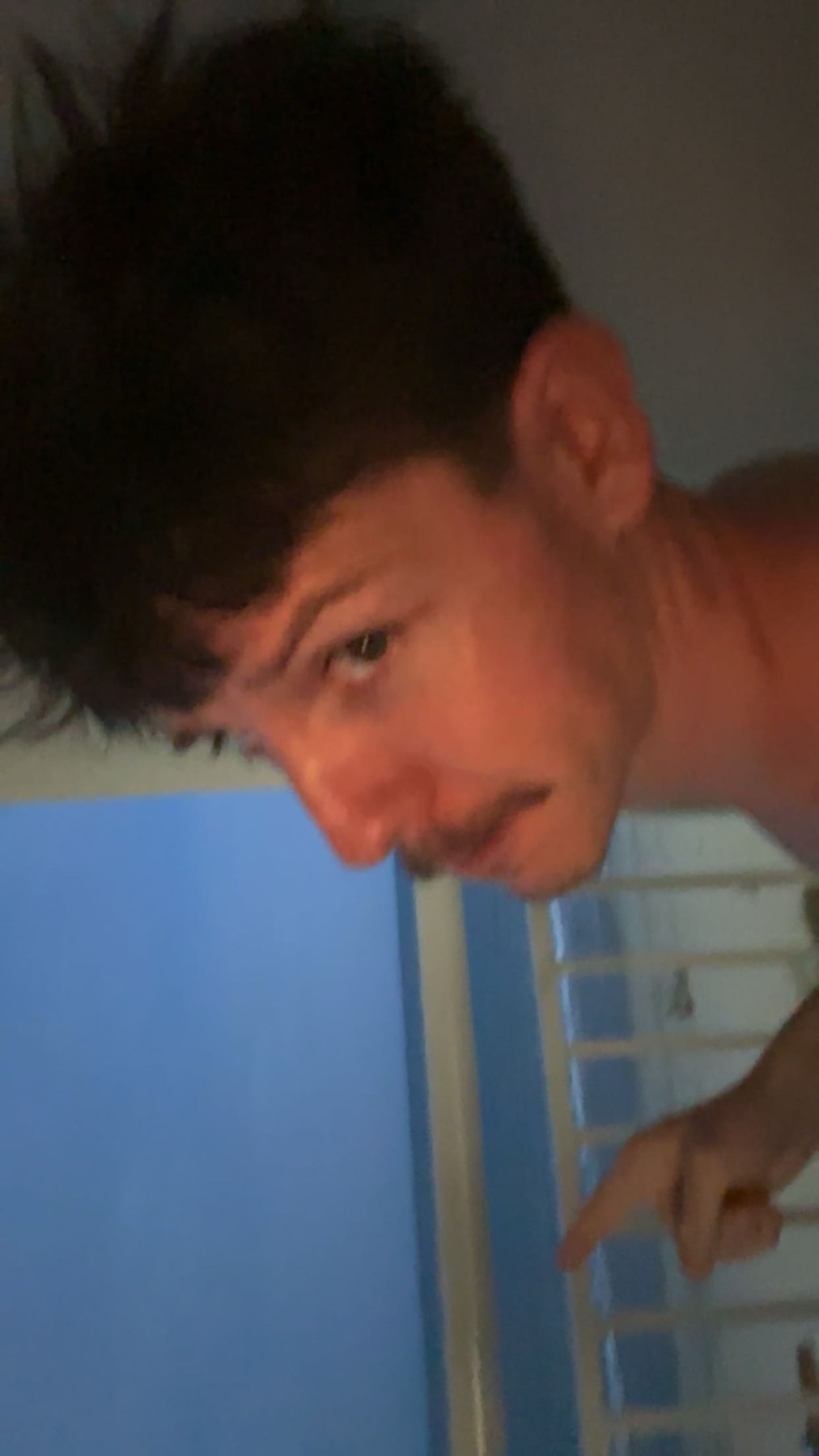 A man with dark, messy hair and a mustache looks down and points to the right. The background is a blurry blue wall and a white slatted surface.