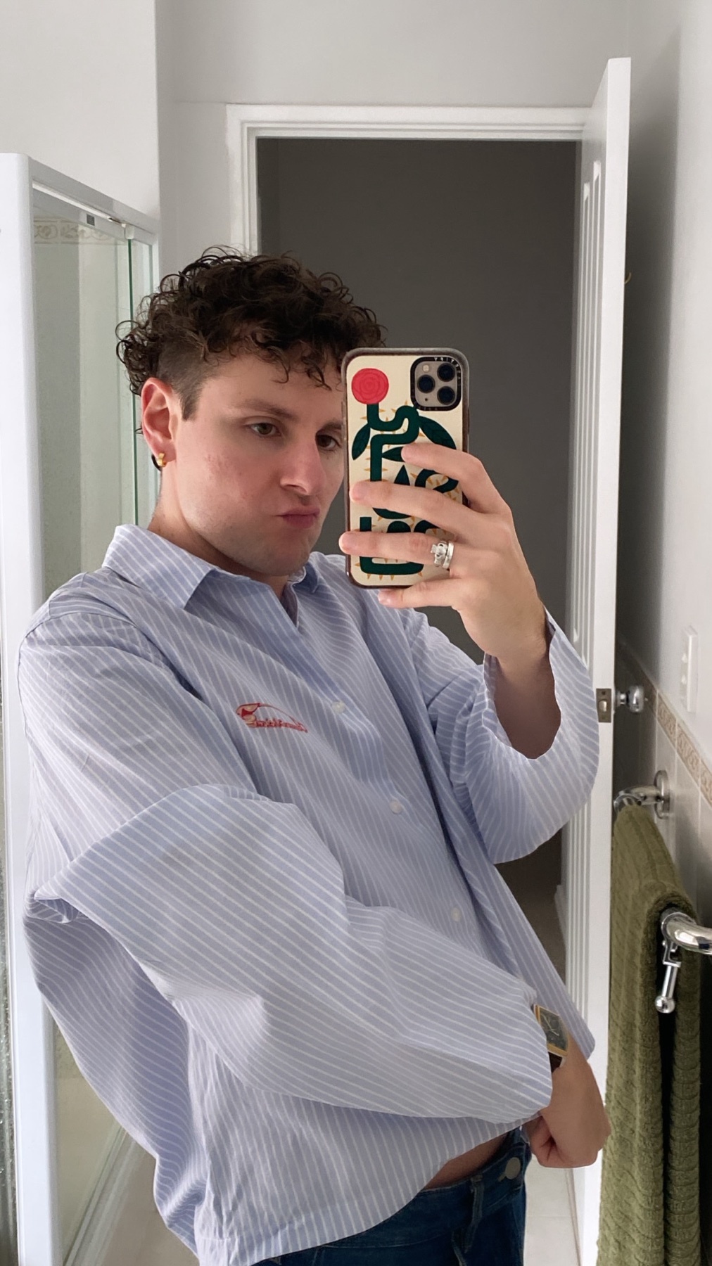 A man with curly hair takes a mirror selfie in a bathroom, wearing a blue and white striped button-down shirt and jeans. He is posing with his phone, which has a colorful, artistic case.