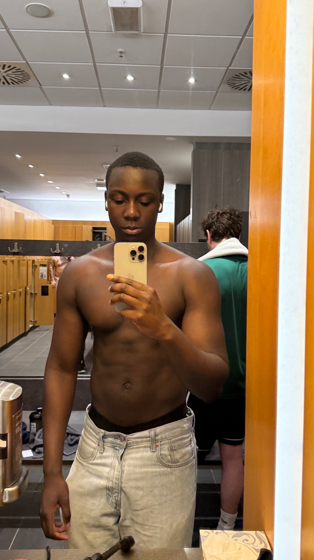 A shirtless Black man takes a mirror selfie in a gym locker room. He is wearing light-wash denim jeans and holding his phone up to take the photo.