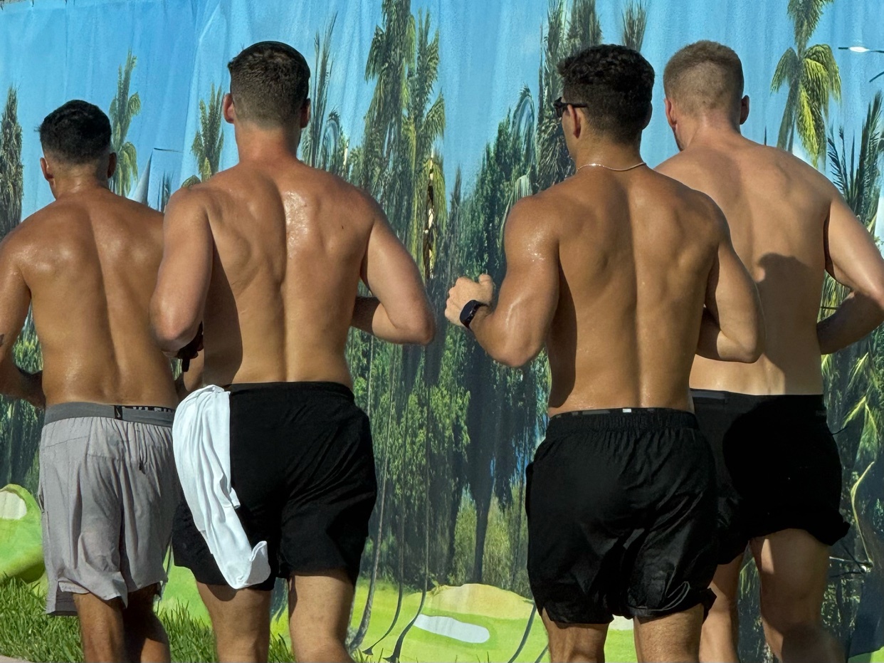Four muscular men are shown from behind, shirtless and sweaty, wearing athletic shorts. They appear to be running or jogging in front of a tropical-themed backdrop.