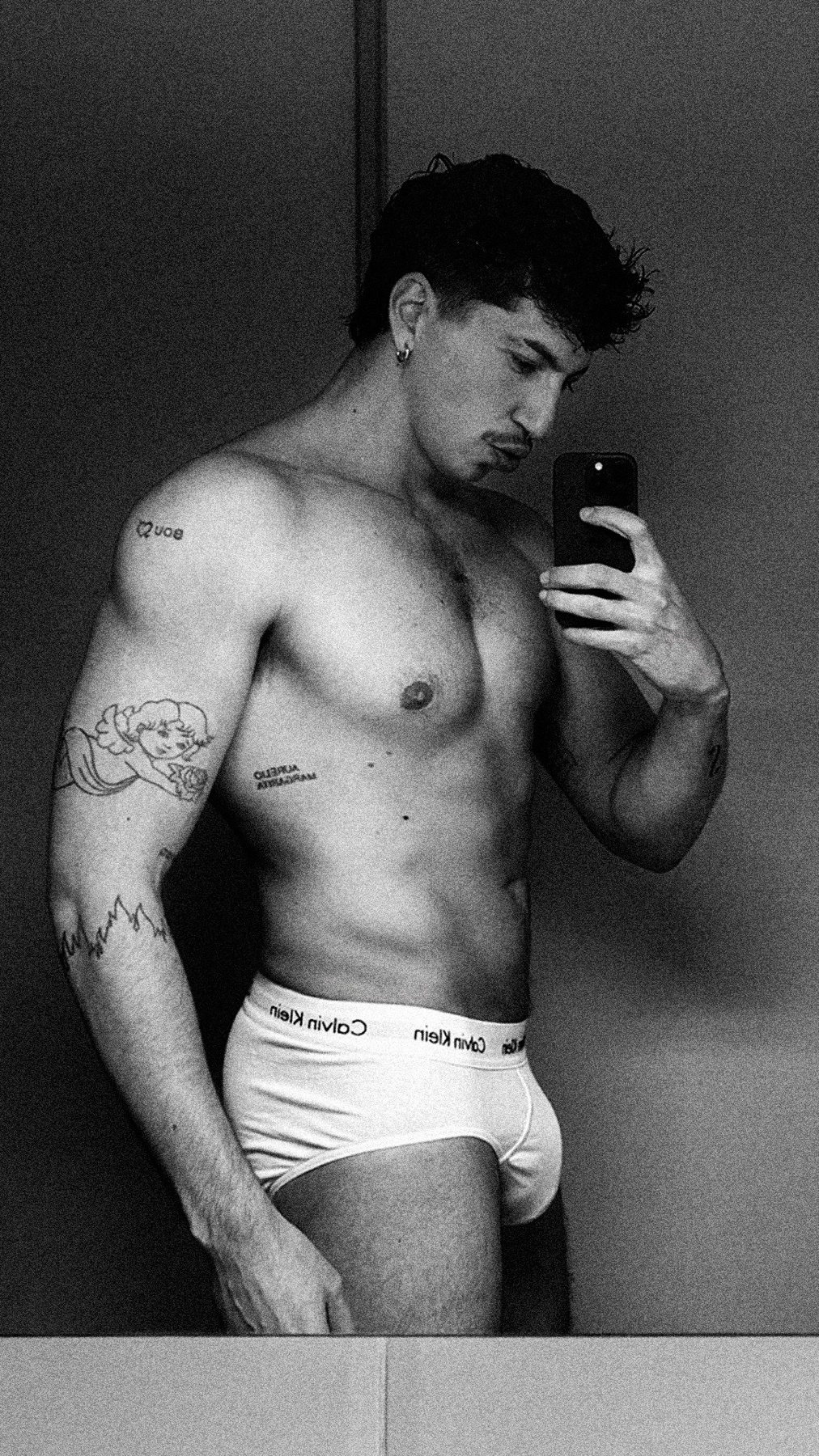 A black and white mirror selfie of a man wearing Calvin Klein underwear. He has tattoos on his arm and torso.