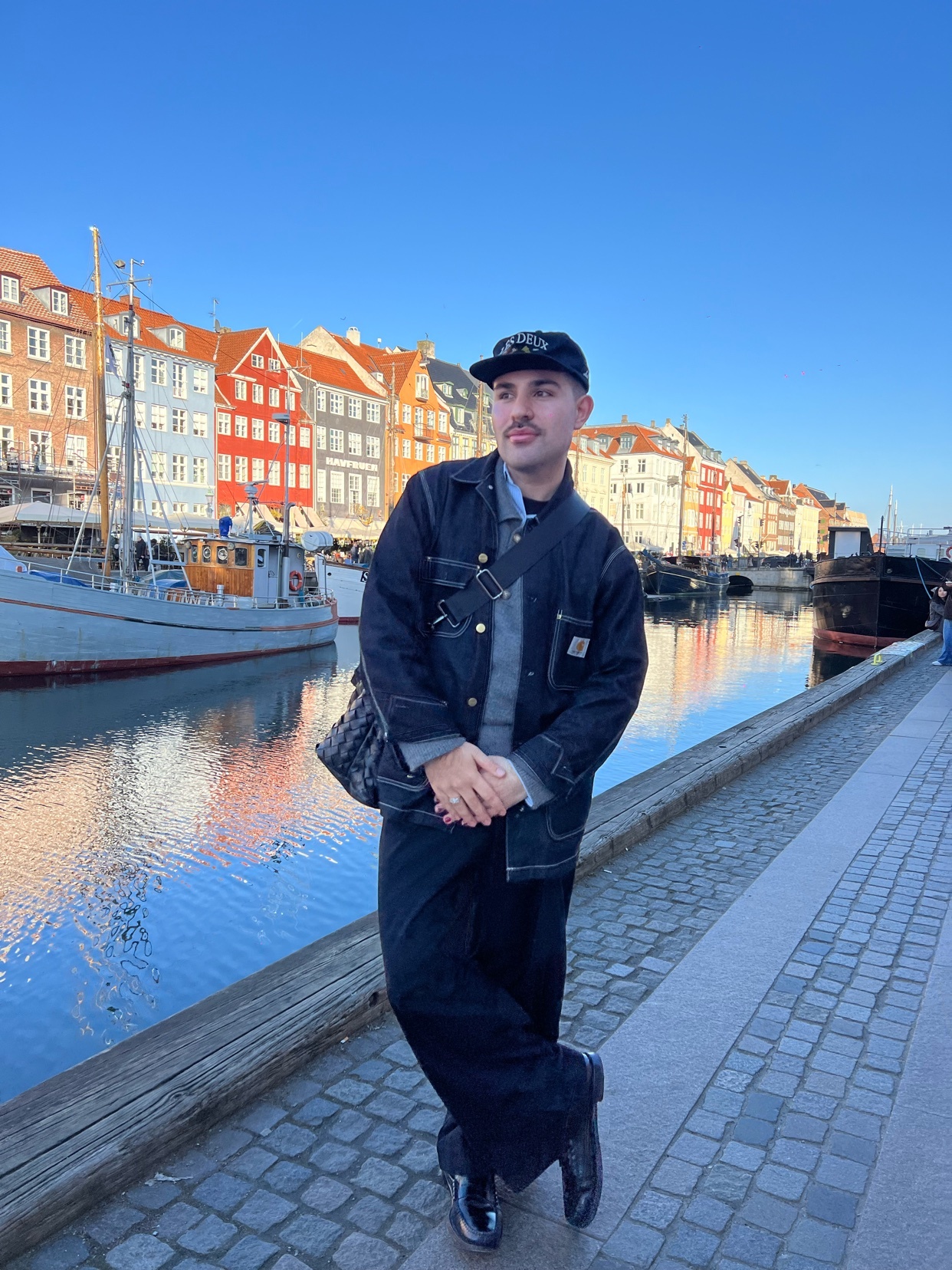 A man poses in front of a canal in Copenhagen. He is wearing a stylish outfit with a jacket and cap.