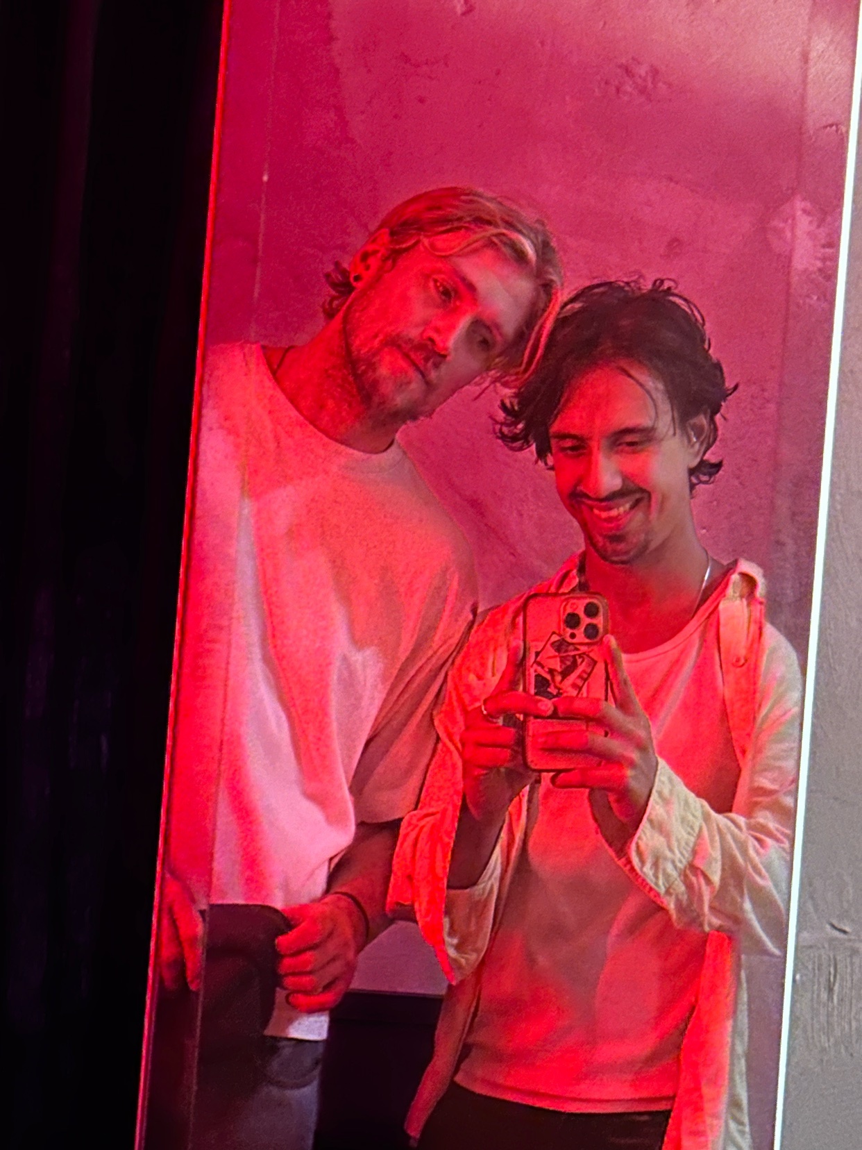 A couple of men take a mirror selfie in a room illuminated by red light. The man on the left has blond hair and a beard, while the man on the right has dark hair and stubble.