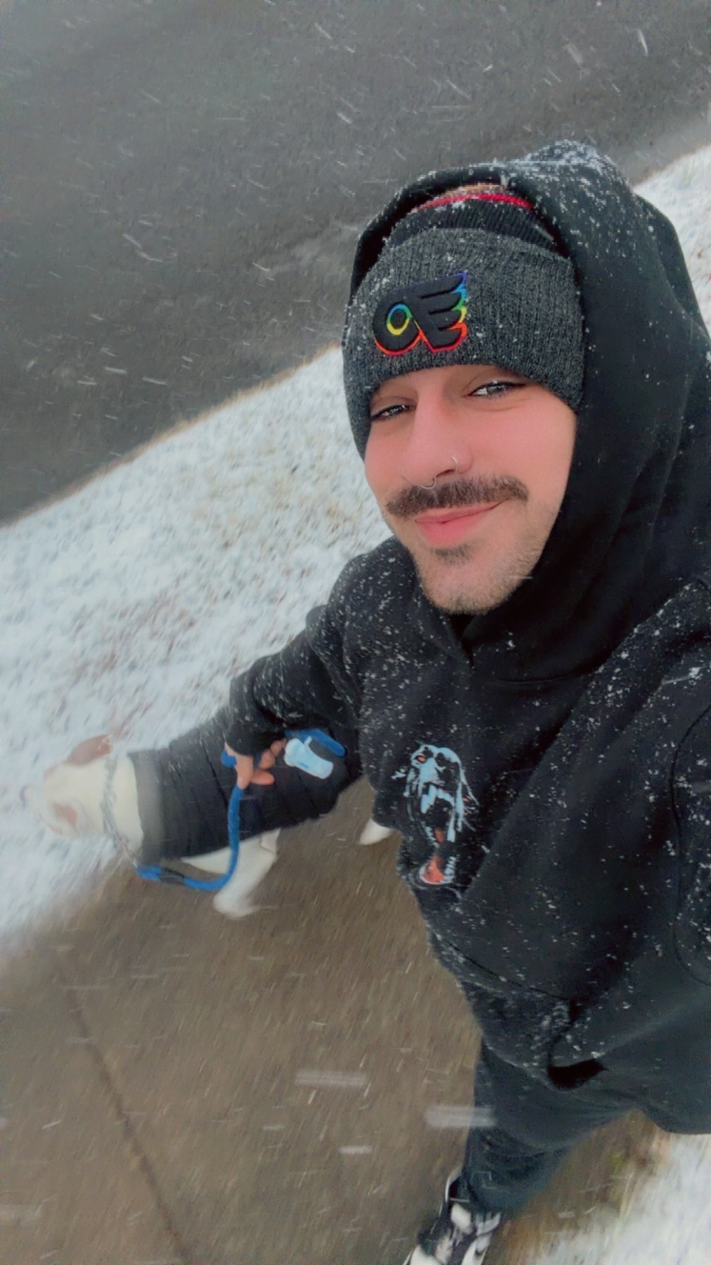 A man takes a selfie while walking his dog in the snow. The man is wearing a beanie and a hoodie.