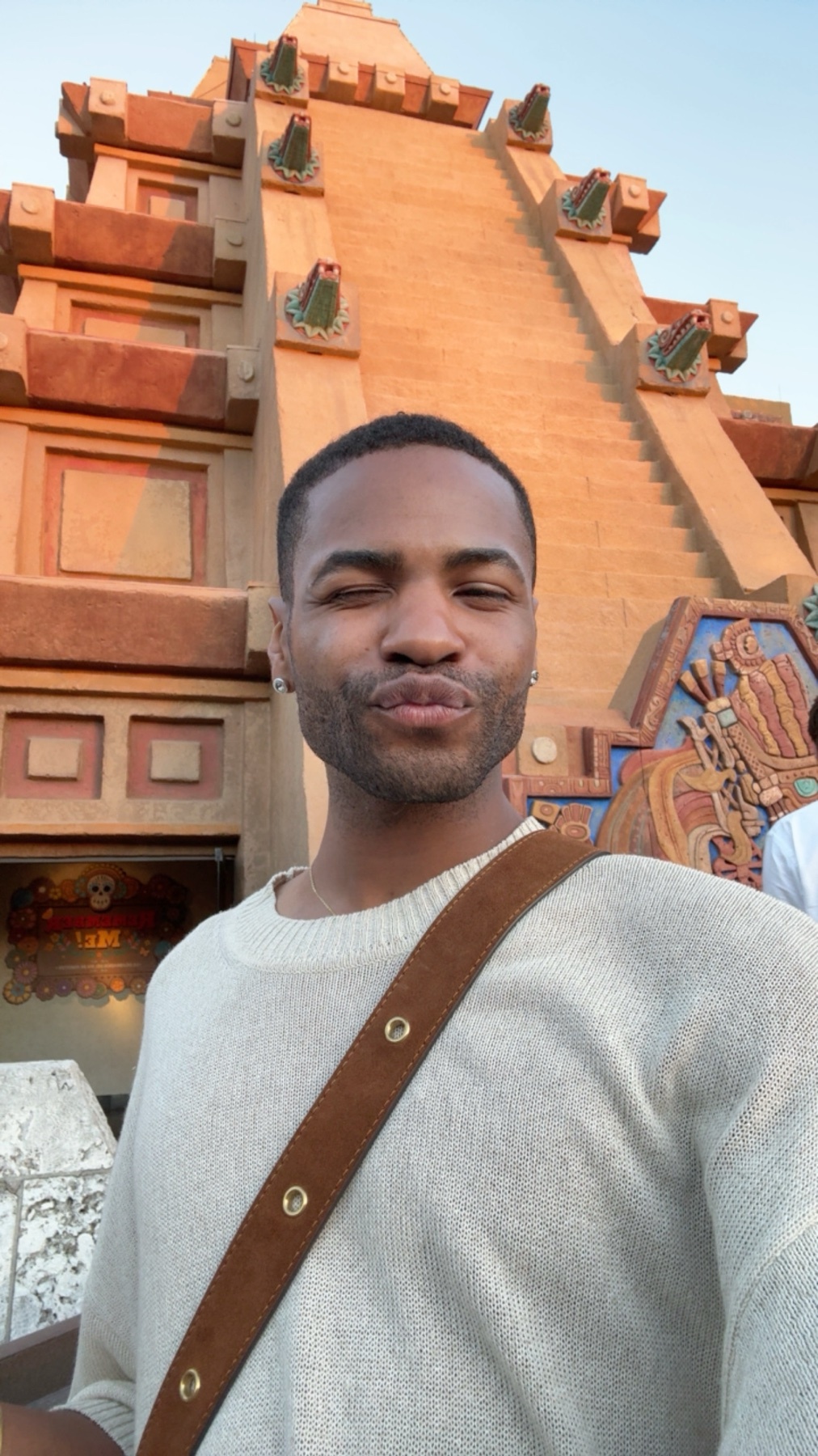 A man poses for a selfie in front of a large, ornate temple structure. The man is wearing a light-colored sweater and a brown crossbody bag, with a playful expression on his face.