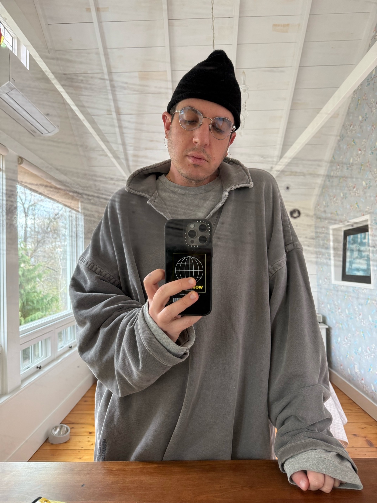 A man takes a mirror selfie, showcasing his outfit. He wears a beanie, glasses, and a stylish oversized hoodie.