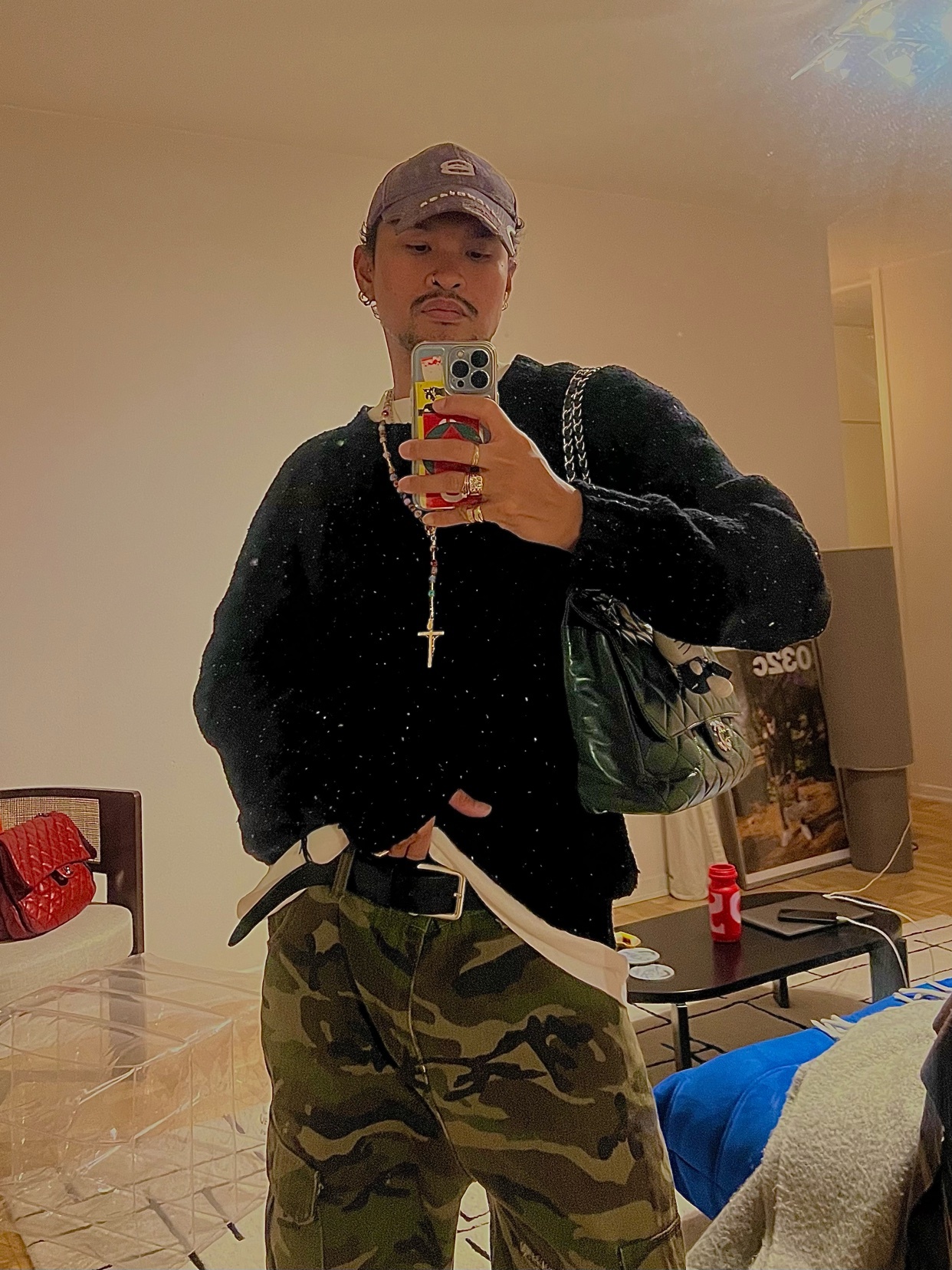 A man takes a mirror selfie, showcasing his outfit. He wears a black sweater, camouflage pants, and a cap, embodying a modern streetwear style.