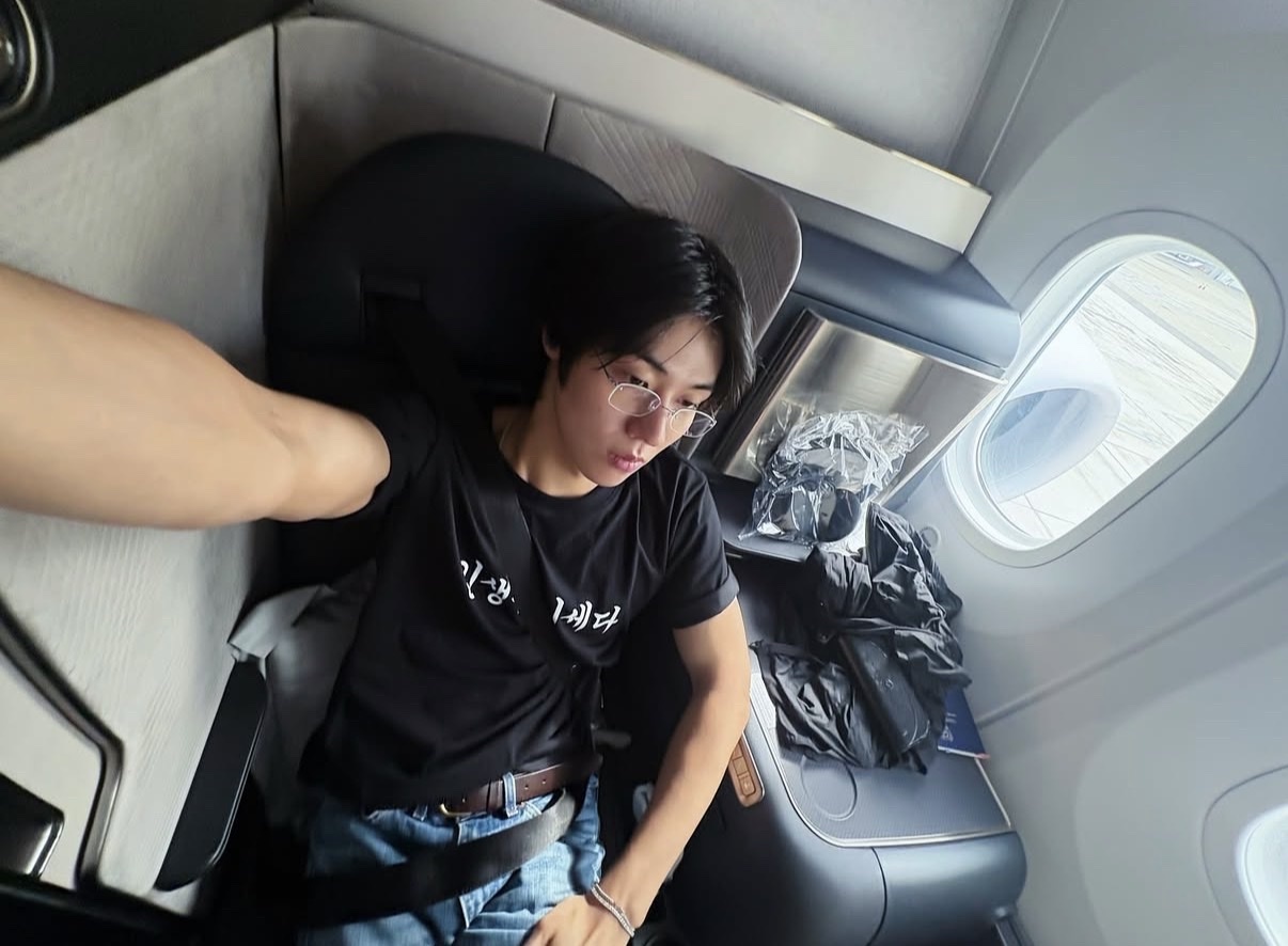 A man takes a selfie while sitting in a first-class airplane seat. He is wearing glasses and a black t-shirt.