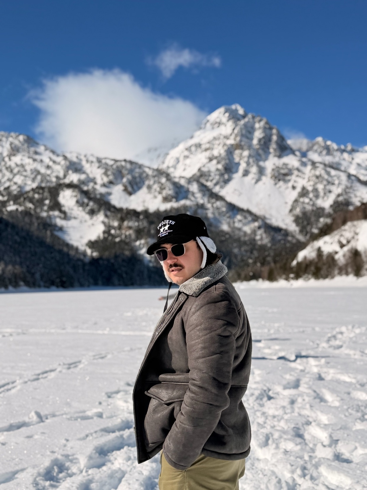 A man poses in front of a snowy mountain range. He is wearing a hat, sunglasses, and a coat.