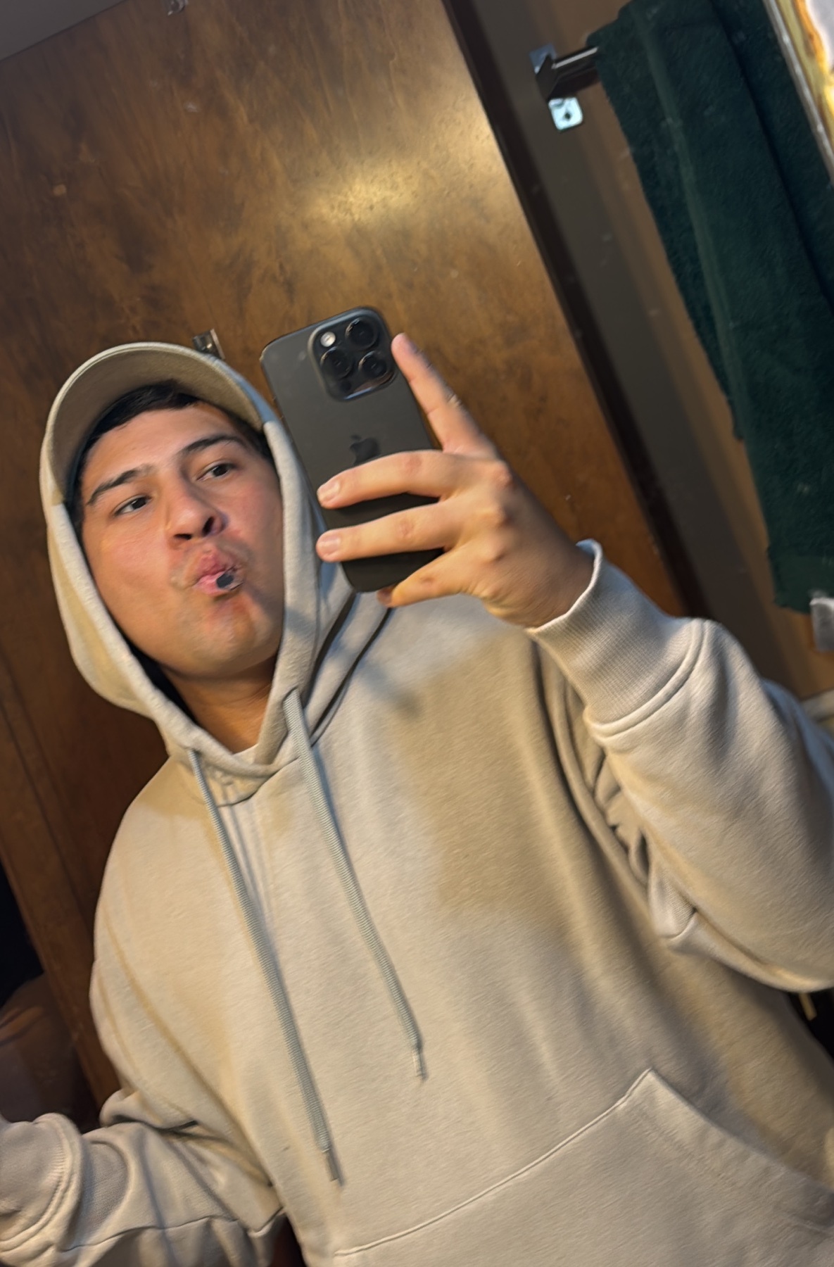 A man takes a selfie in a mirror, wearing a hoodie and cap. He is making a kissing face.