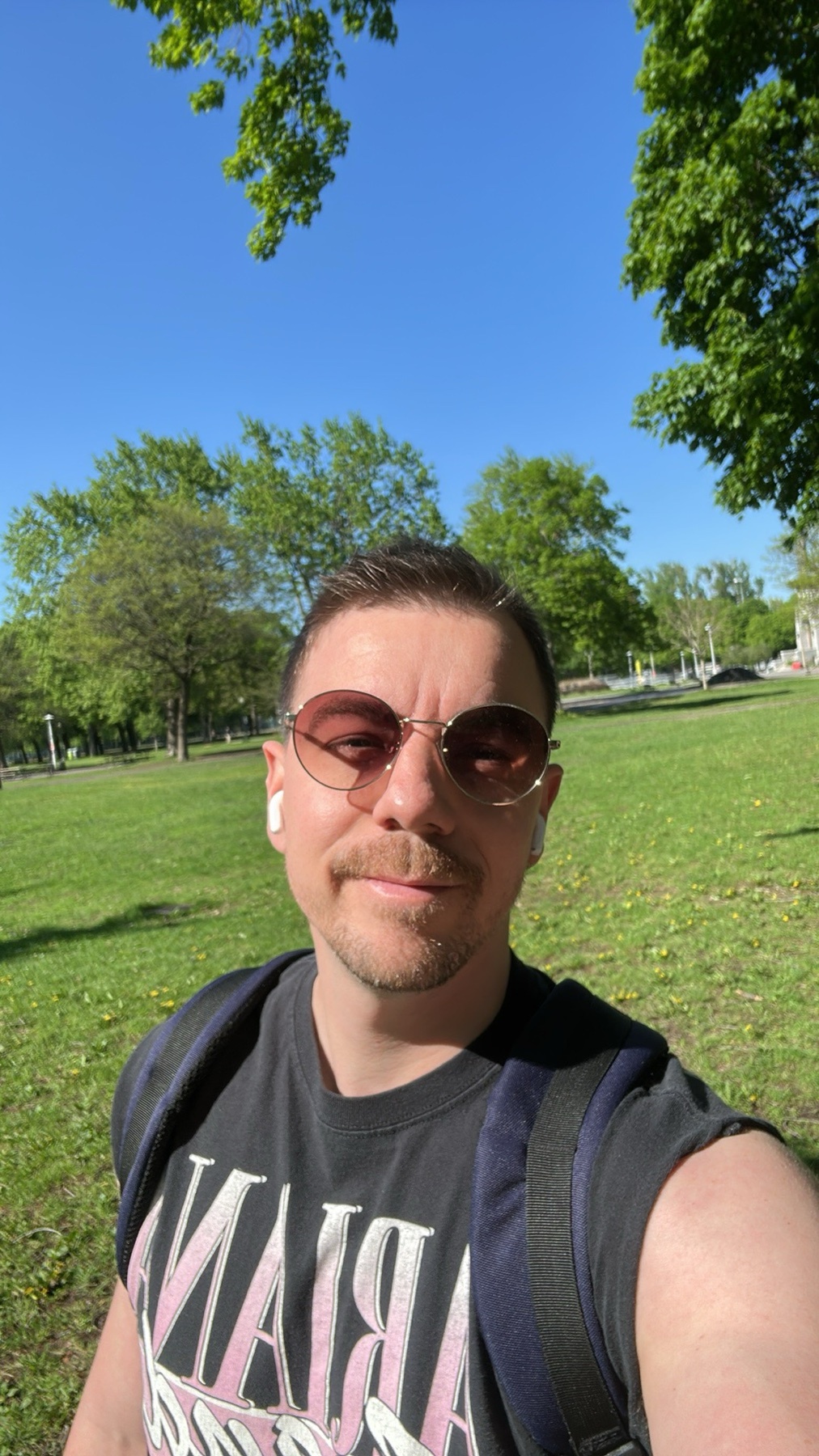 A man wearing sunglasses and earbuds takes a selfie in a park on a sunny day. He is wearing a t-shirt and a backpack, with green grass and trees in the background.