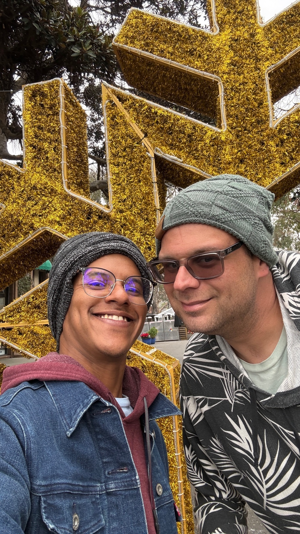 A smiling couple poses for a selfie in front of a large, golden snowflake decoration. They are bundled up in winter attire, suggesting a festive holiday outing.