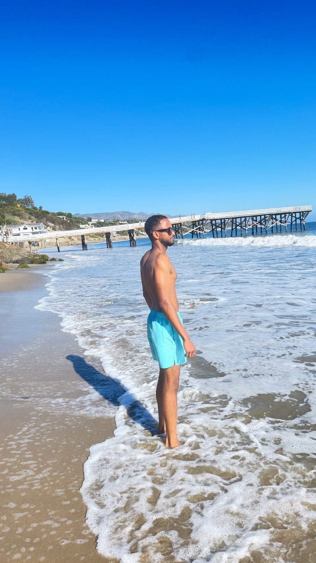 A man stands at the edge of the ocean, wearing blue swim trunks and sunglasses. He looks out towards a distant pier under a clear blue sky.