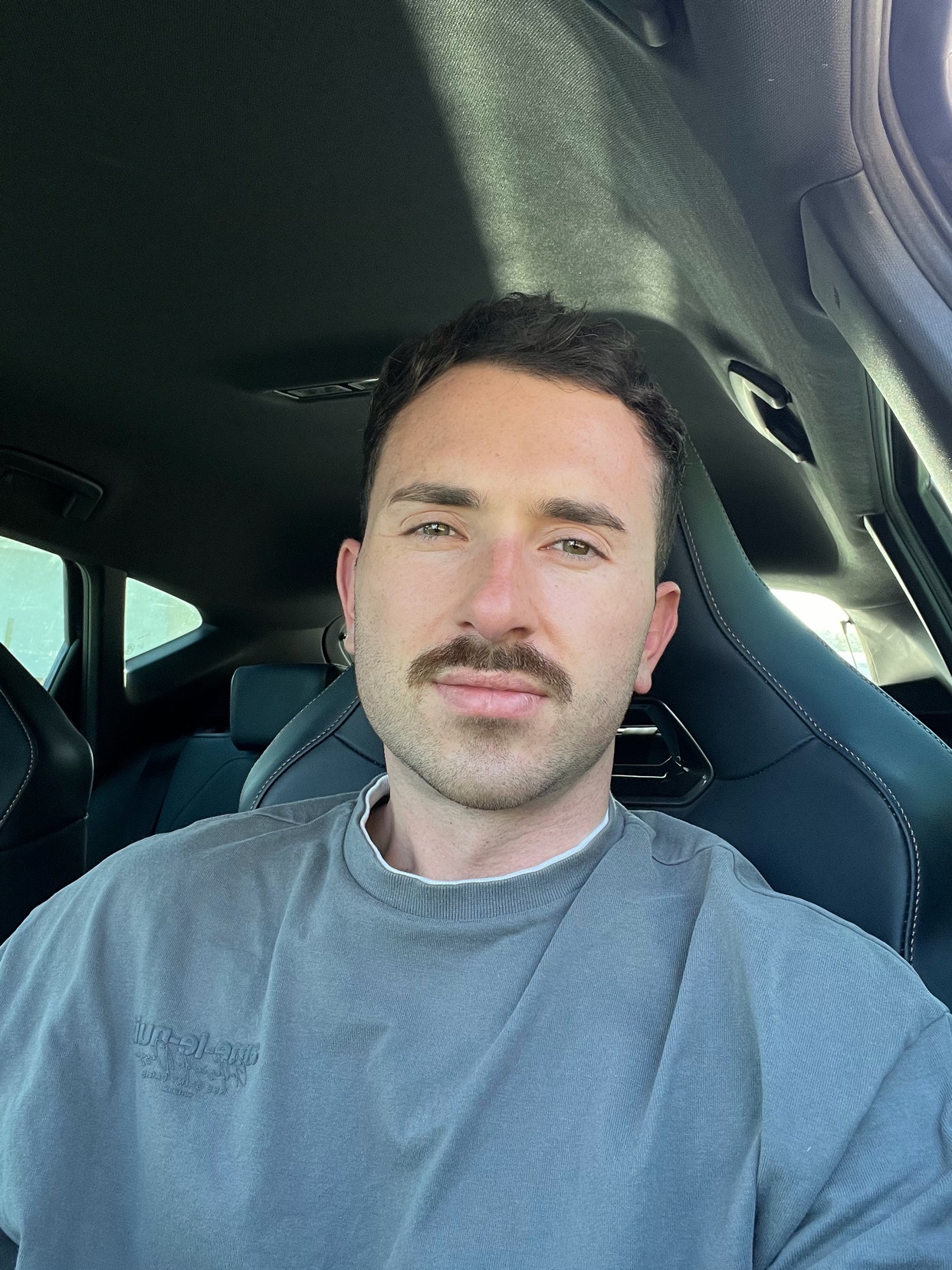A man takes a selfie while sitting in a car. He has dark hair and a mustache.