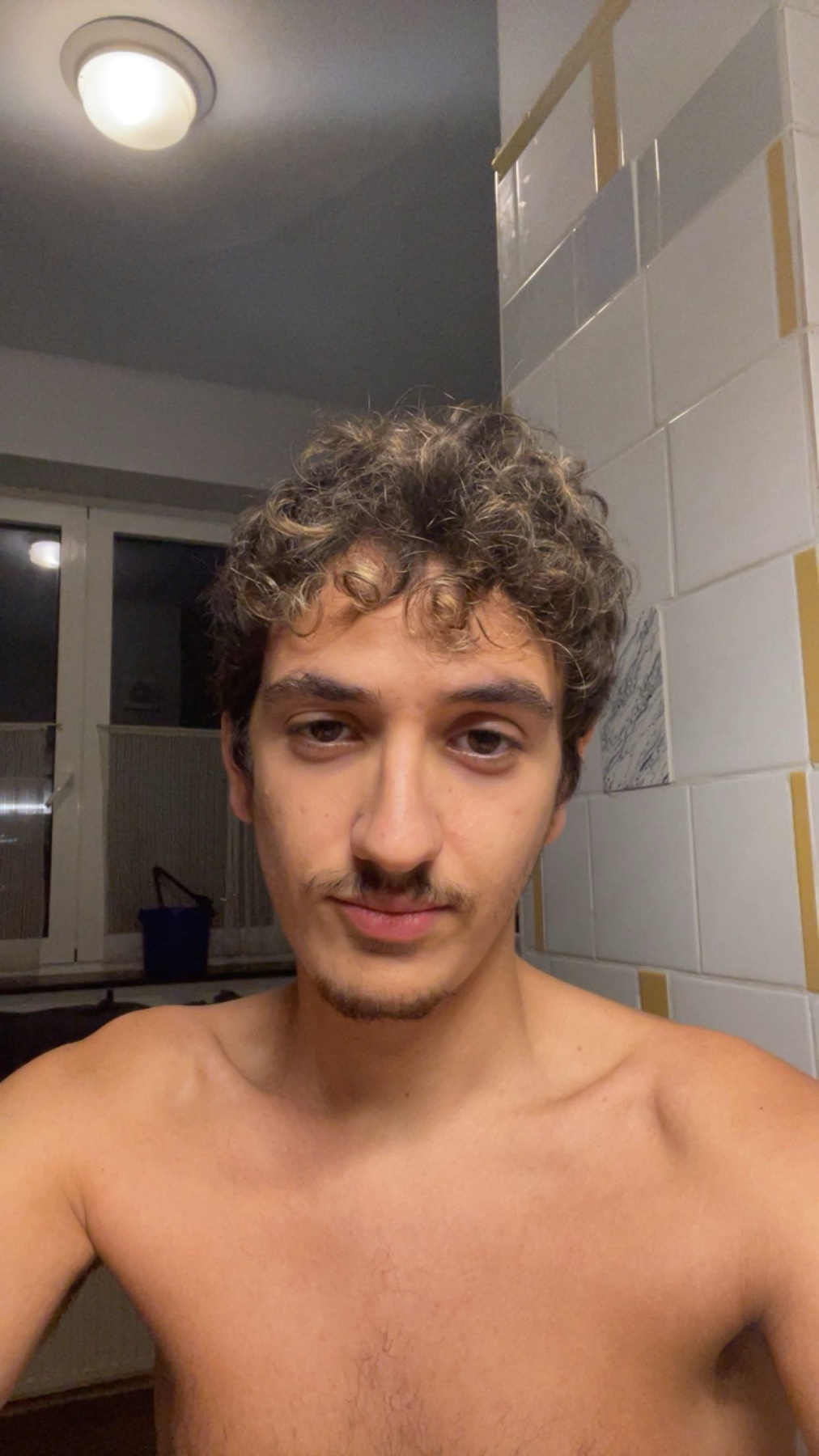 A selfie of a man with curly hair and a mustache. He is shirtless.