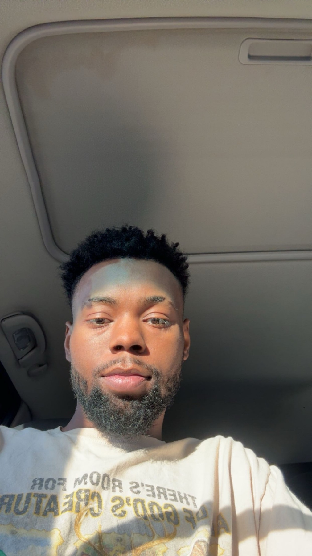 A man takes a selfie in a car, bathed in sunlight. He has dark hair and stubble.