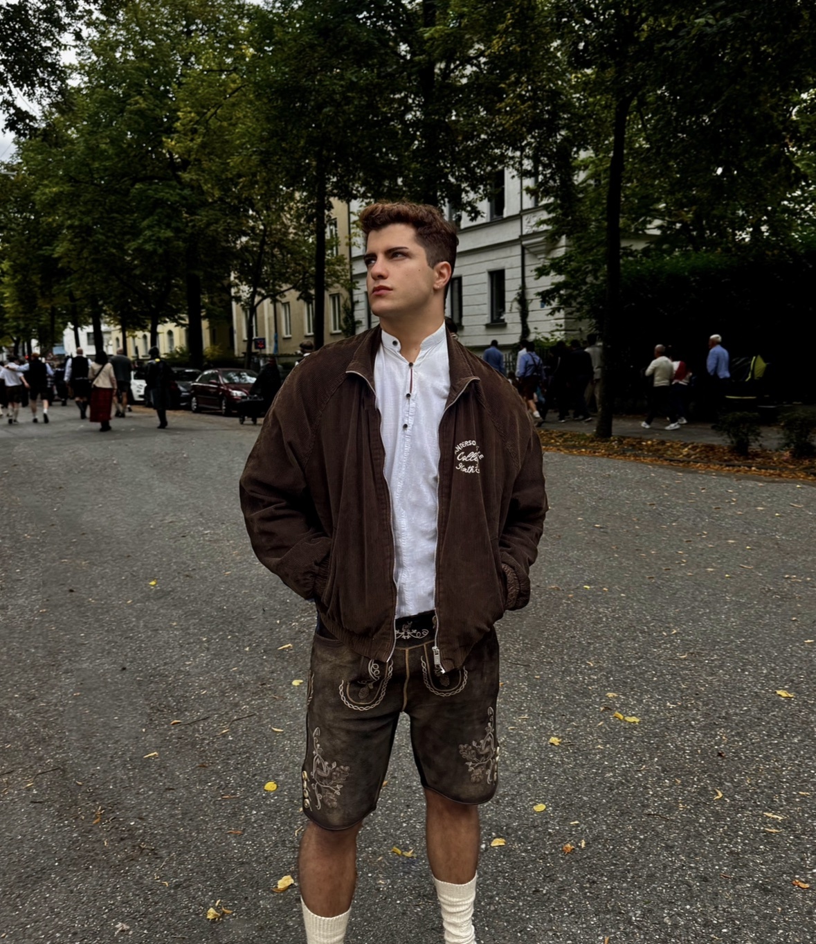 A young man poses outdoors wearing traditional Bavarian lederhosen and a white dirndl shirt. He is dressed in a brown jacket and white socks, standing on a paved street lined with trees.