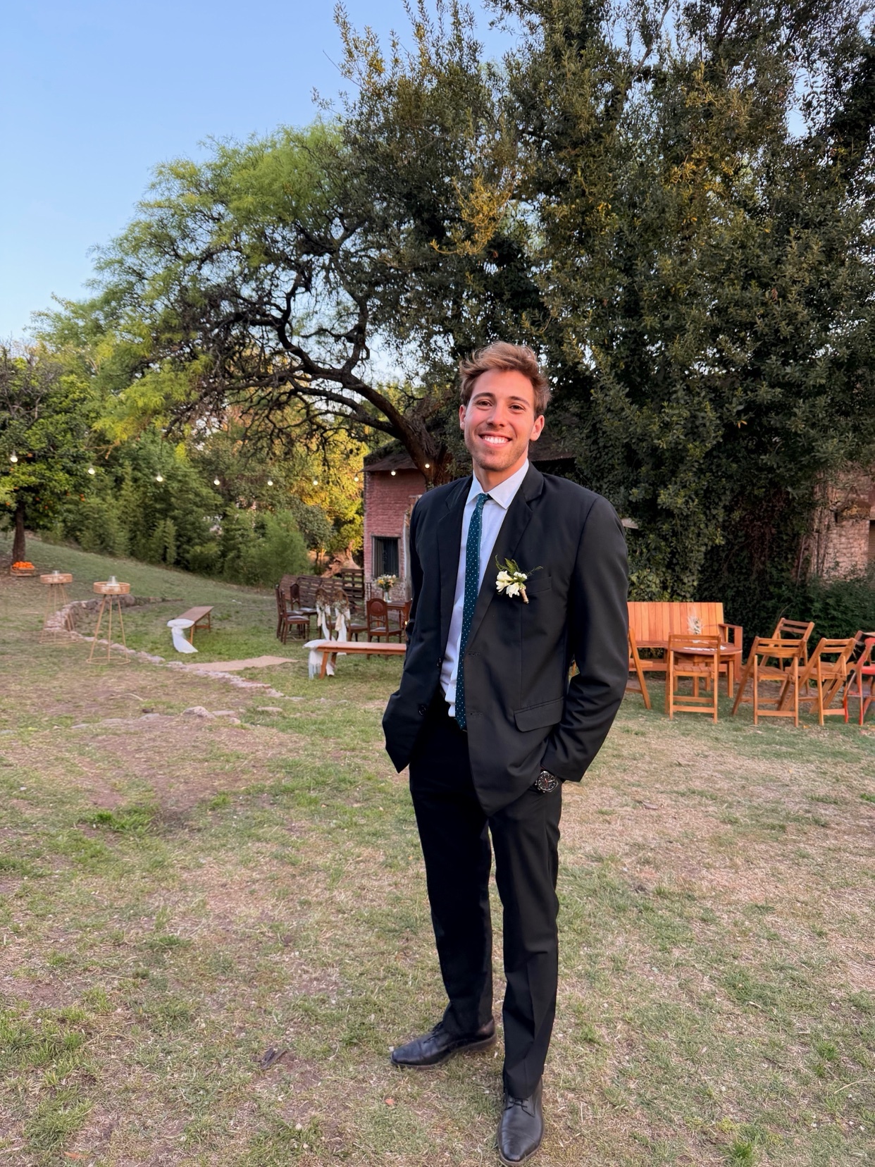 A man in a black suit, white shirt, and blue tie smiles as he stands in a grassy outdoor setting with trees and chairs in the background.