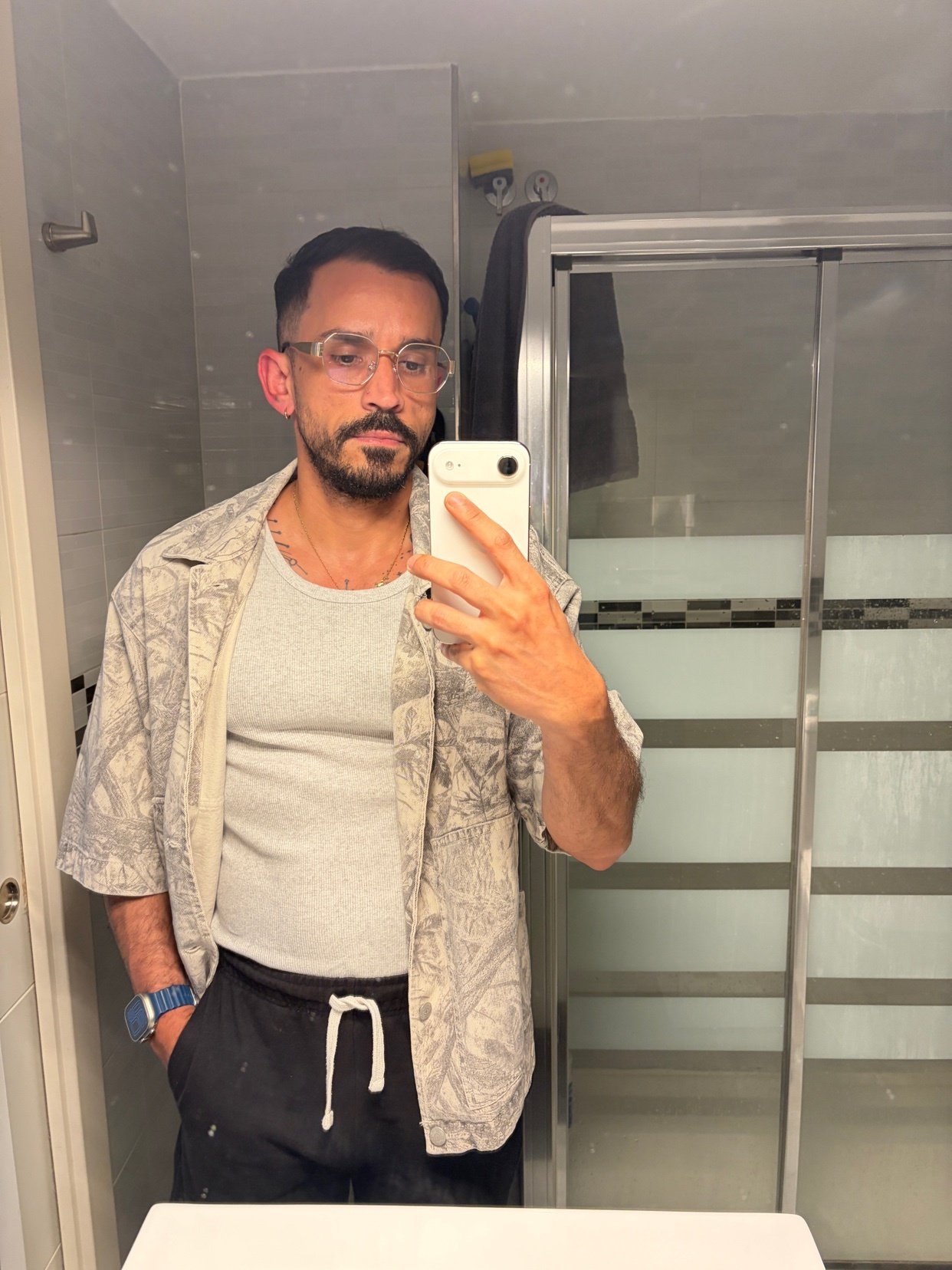 A man with glasses and a beard takes a selfie in a bathroom mirror. He is wearing a grey tank top and an open, short-sleeved patterned shirt.