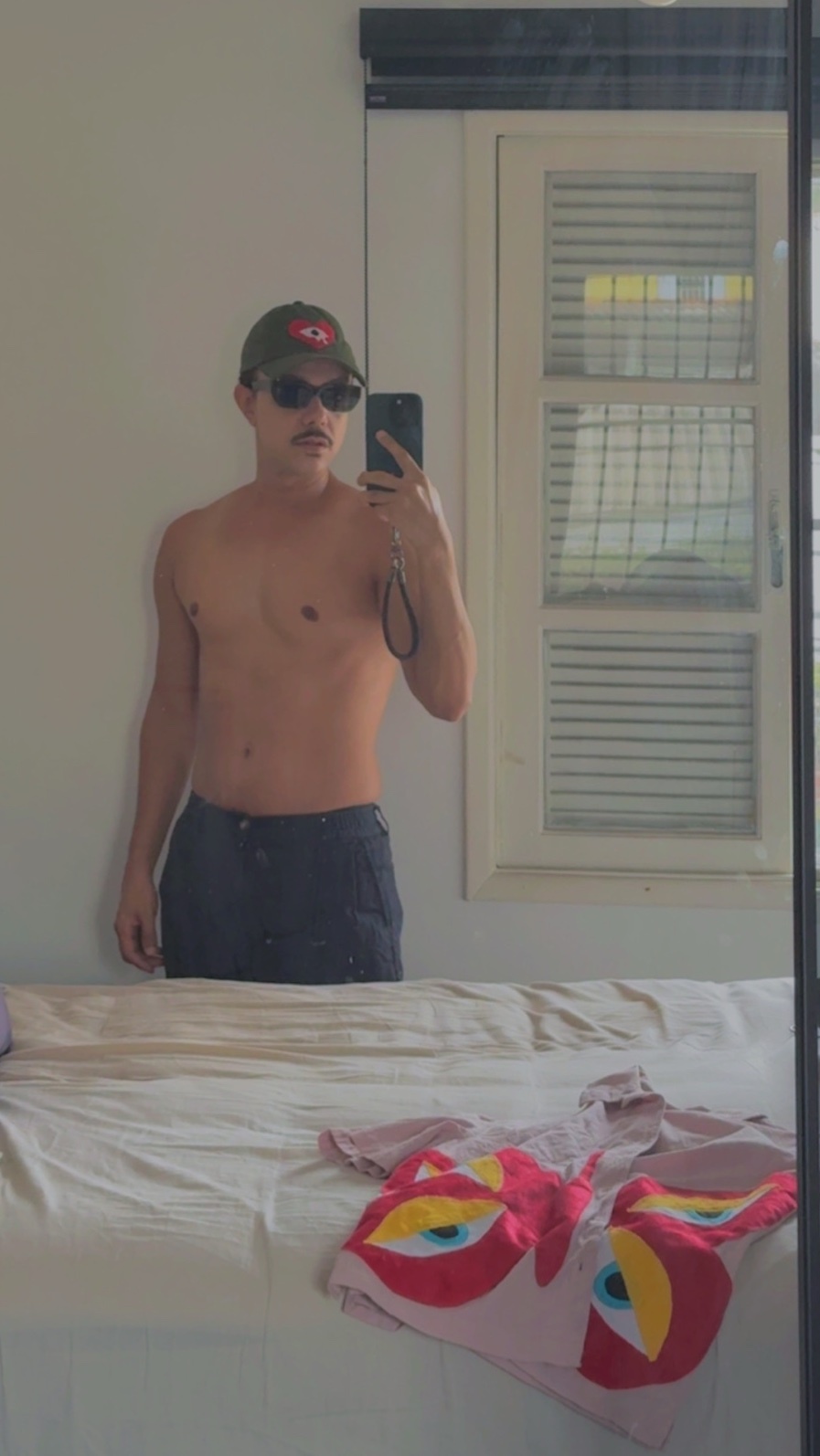 A man in sunglasses and a baseball cap takes a mirror selfie. He is shirtless and wearing dark pants.