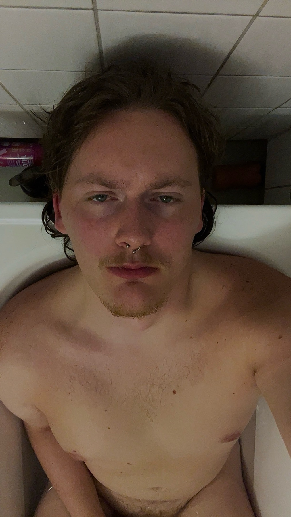 A shirtless man sits in a bathtub, looking directly at the camera. The photo is a selfie, with the man's face and upper body in focus.
