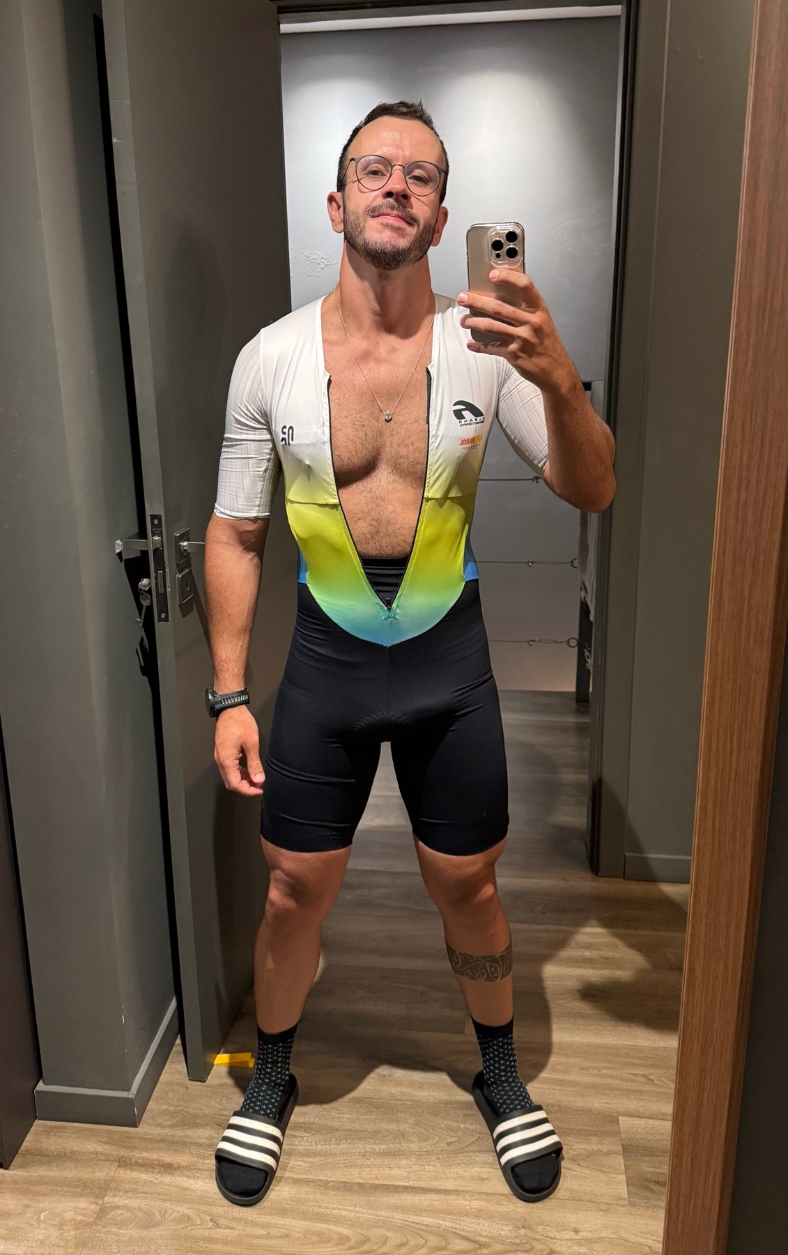 A man in a cycling outfit takes a selfie in a locker room. He is wearing glasses and has tattoos on his legs.