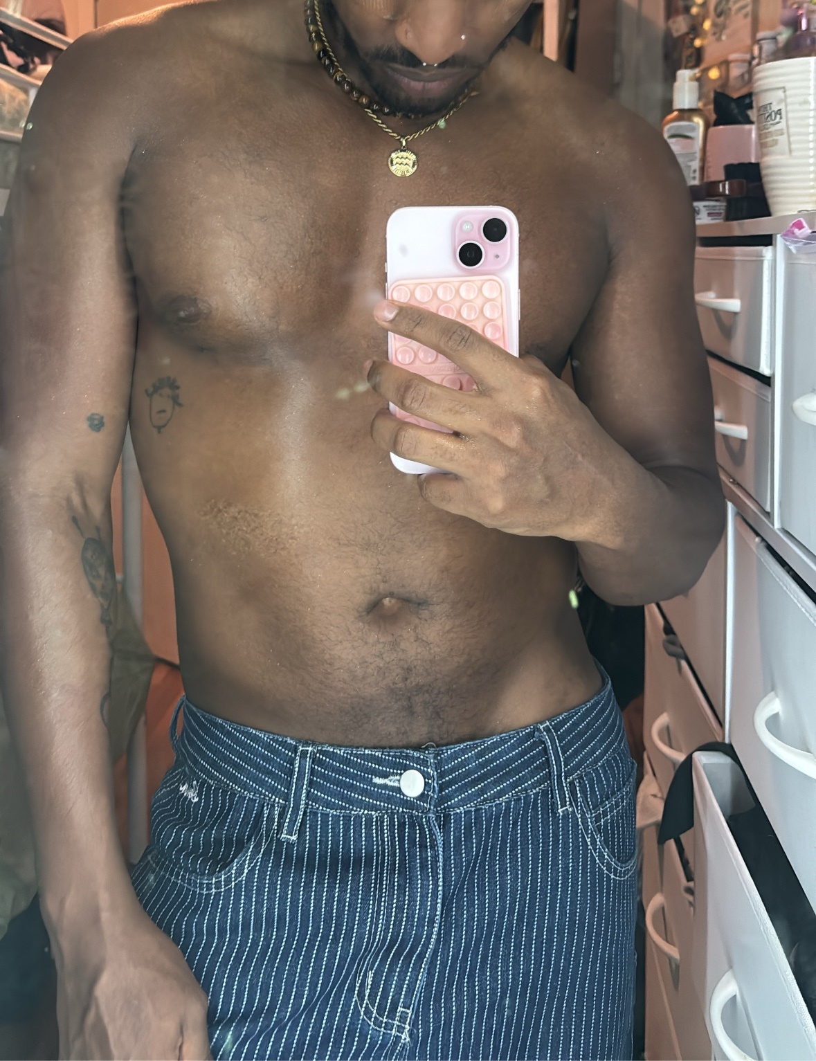 A man takes a selfie in a mirror, showing off his bare chest and abs. He is wearing striped pants and a necklace.