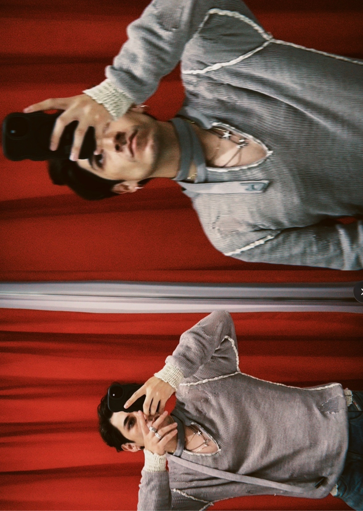 A man takes a selfie in front of a red curtain, holding a camera up to his face. He is wearing a grey, distressed tunic with lace-up detailing.
