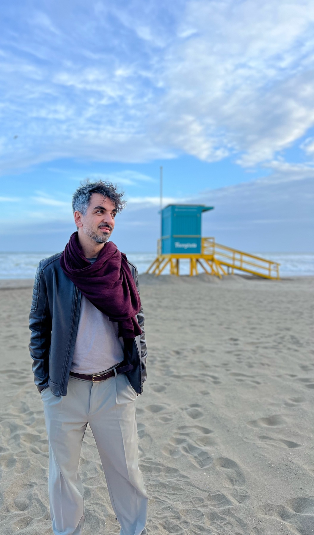 A man poses on a beach, wearing a leather jacket and scarf. A lifeguard stand is visible in the background.