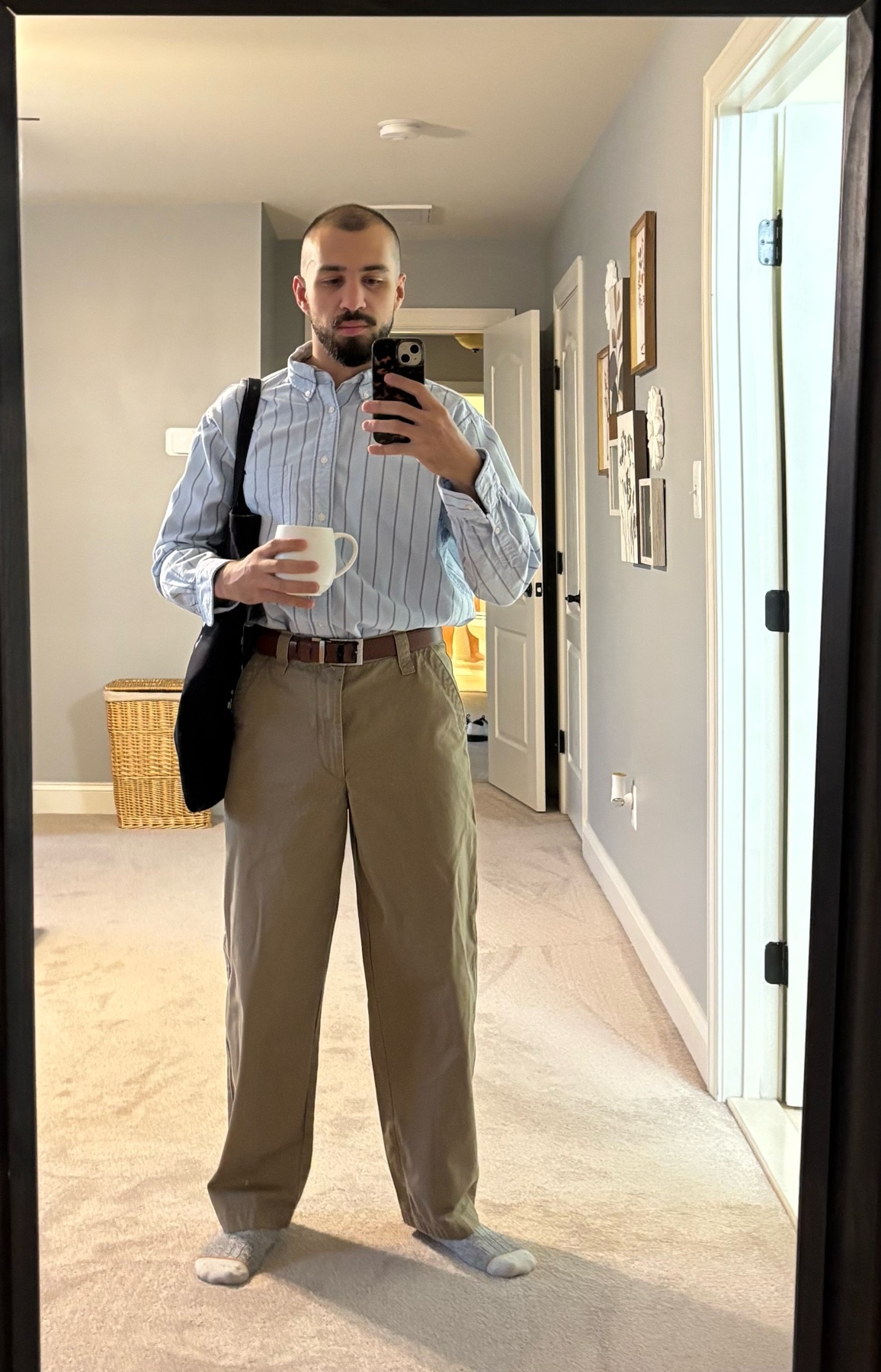 A man takes a mirror selfie, holding a mug and a phone. He is dressed in a striped shirt and khaki pants.