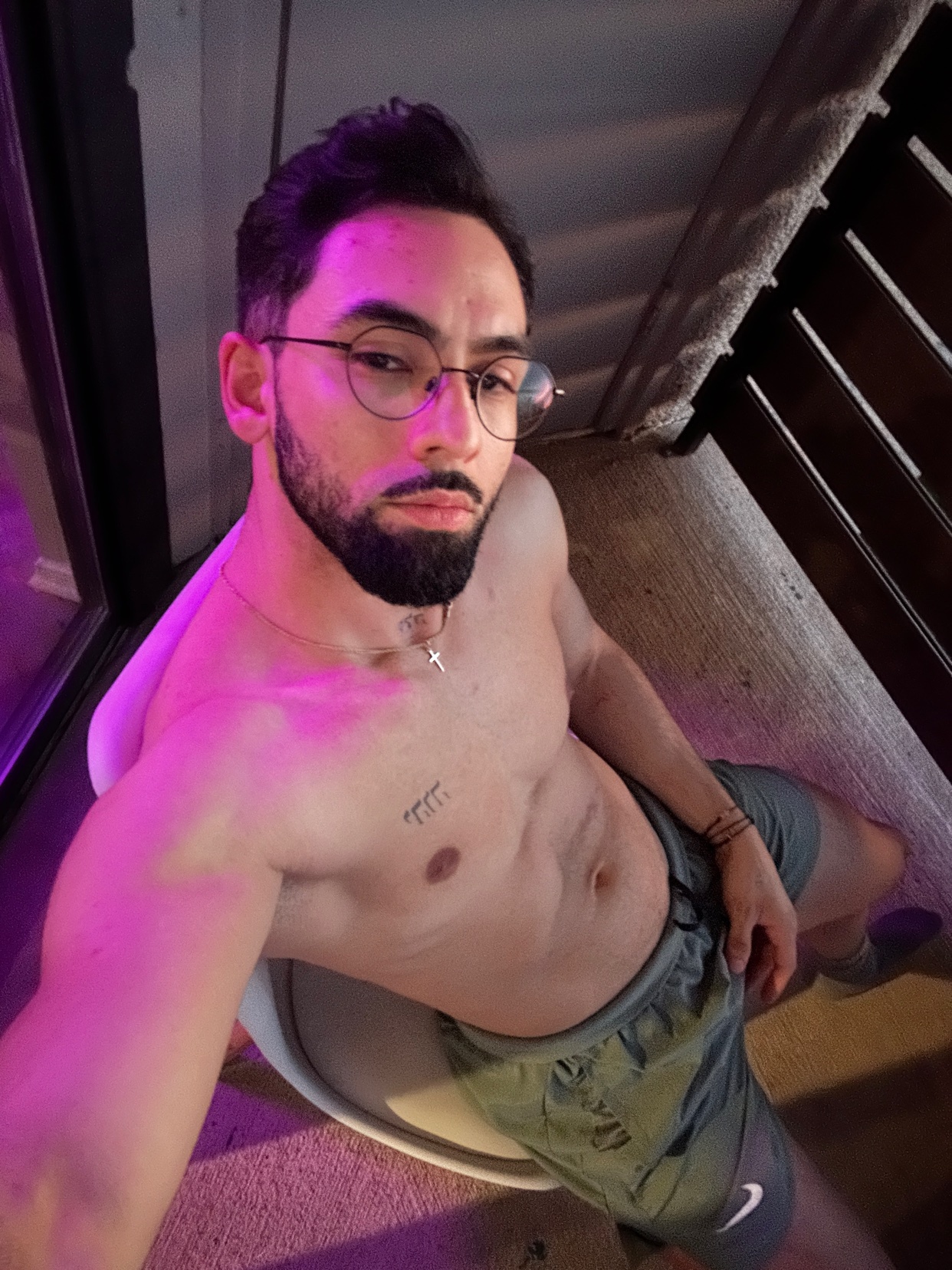 A man with a beard and glasses takes a selfie, showing off his athletic physique. He is shirtless and wearing athletic shorts.