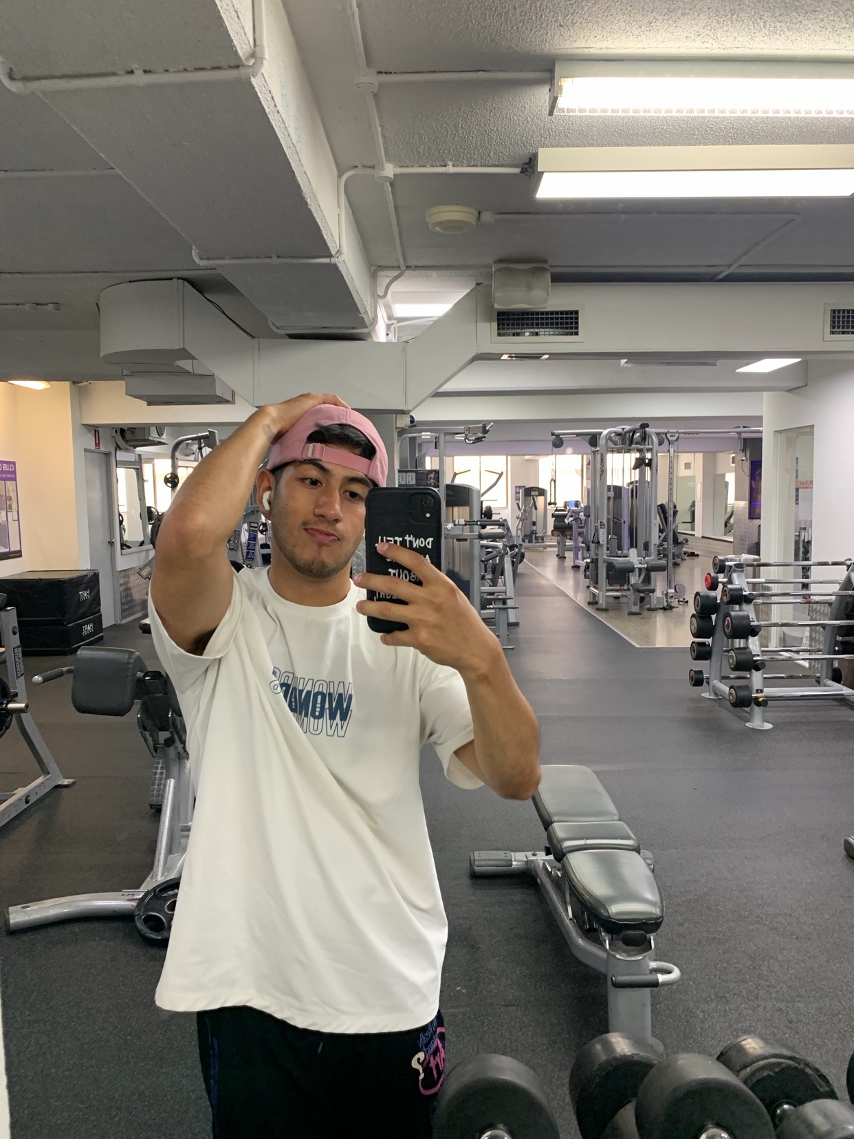 A man takes a mirror selfie at the gym. He is wearing a t-shirt and a cap, and the gym equipment is visible in the background.