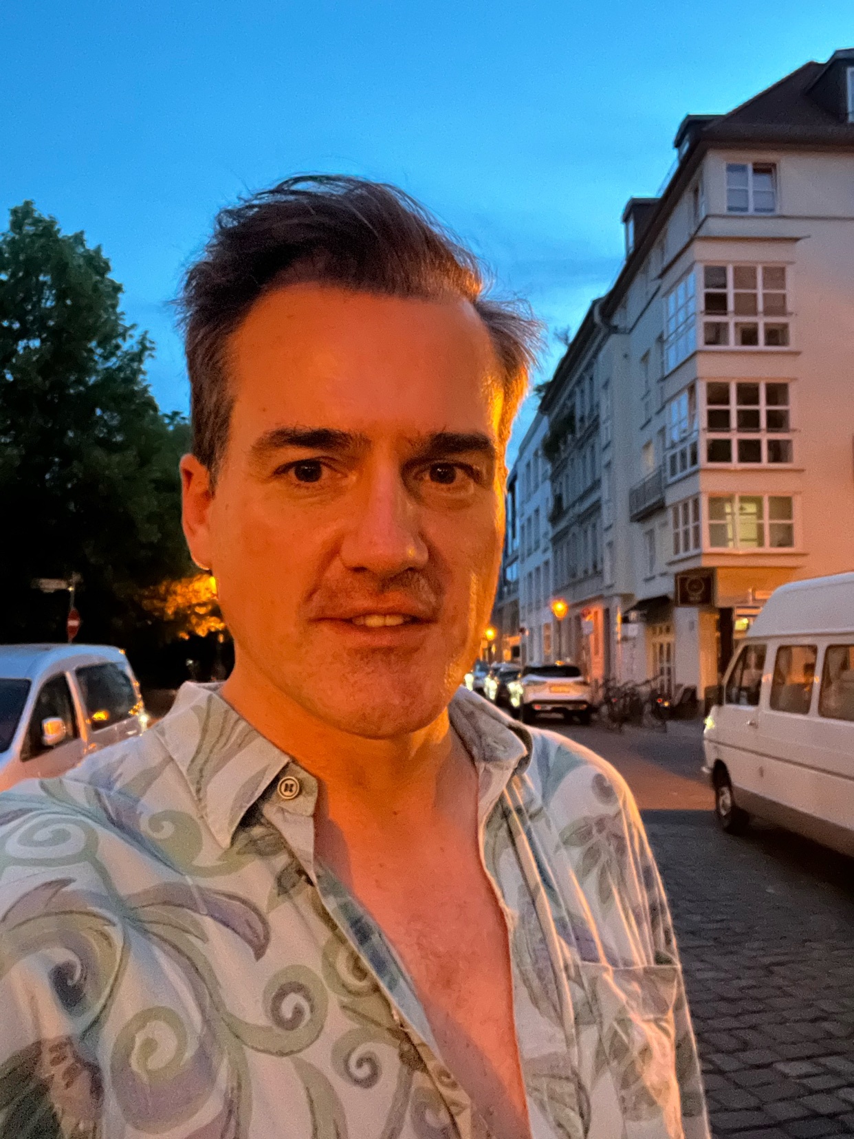 A man takes a selfie outdoors at dusk. He is shirtless and standing in front of a street with buildings.
