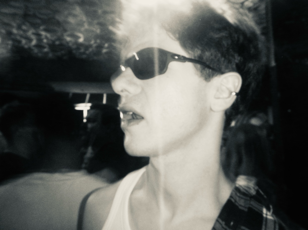 A black and white, close-up, profile shot of a young man wearing sunglasses and a white tank top. He has a flannel shirt draped over his shoulders and is wearing an earring in his left ear.